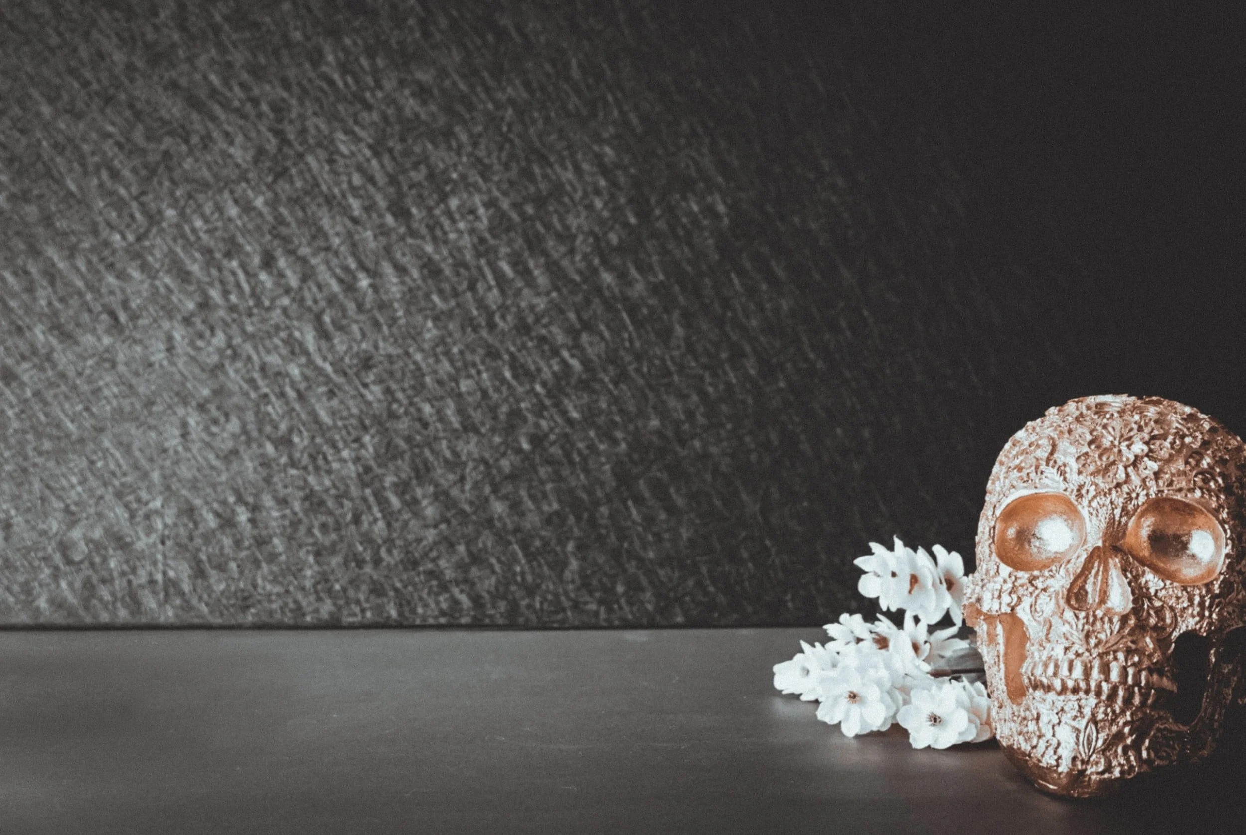 Decorative skull with floral pattern and metallic finish, placed on a dark surface next to white flowers.