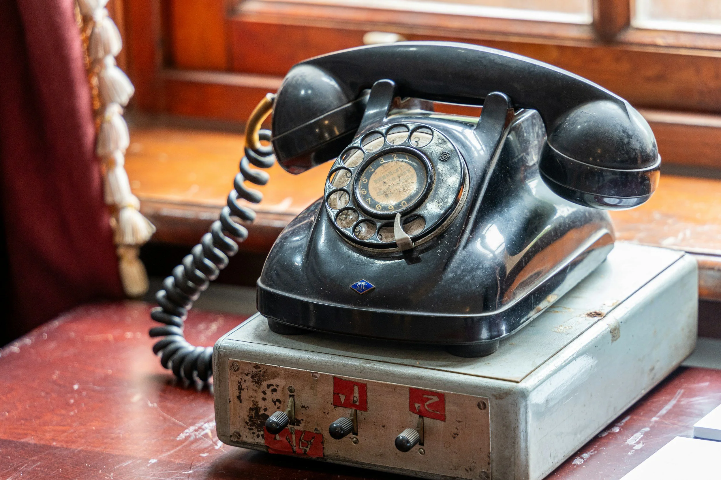 An old black rotary phone placed on a metal base with numbered switches, sitting on a wooden surface near a window.