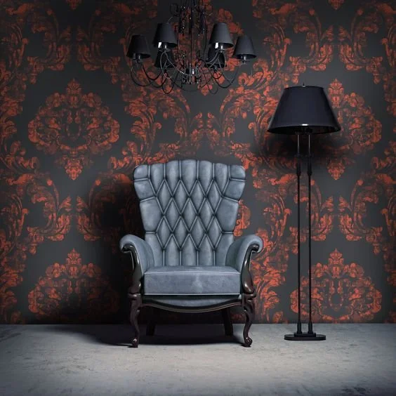 A gray tufted armchair with wooden legs, black chandelier above, and a black floor lamp beside it, set against a red and black patterned wallpaper.