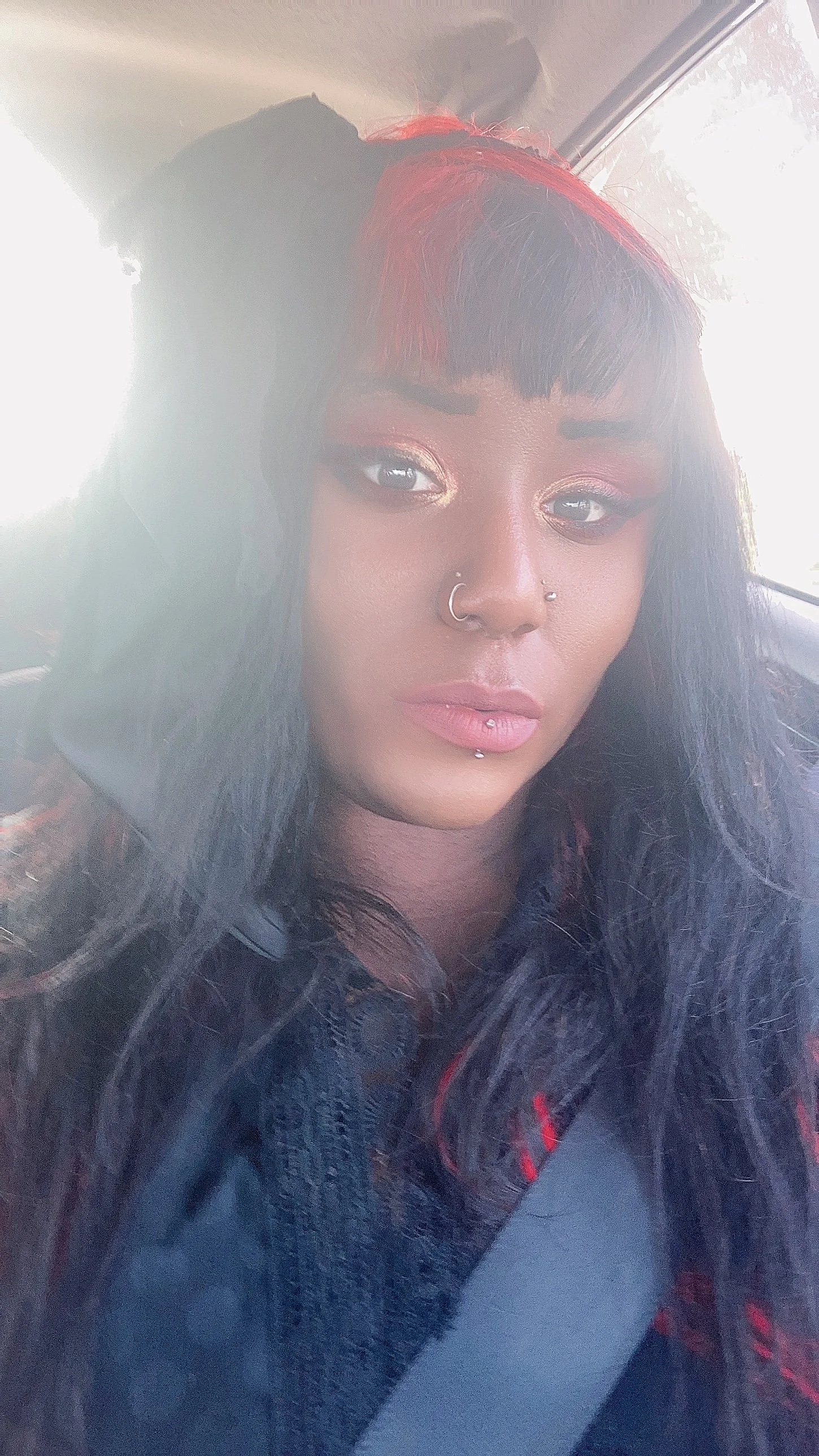 A young woman with long dark hair and red highlights, wearing makeup, jewelry including nose and lip piercings, and a denim jacket, sitting inside a car.