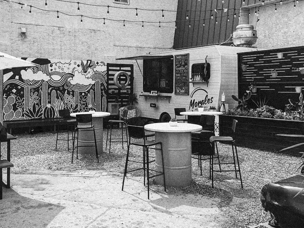 A small outdoor seating area with high tables and chairs, a mural with cloud and plant designs, string lights overhead, and a food truck with the name 'Morales' on it.