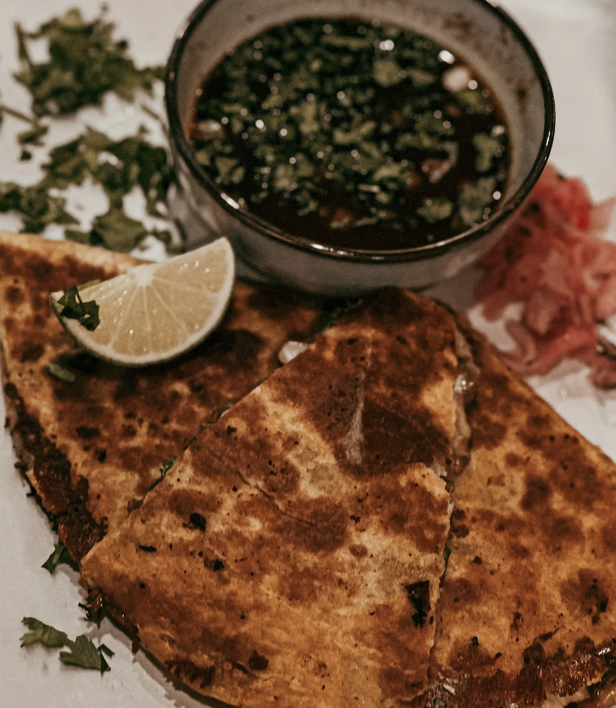 Grilled sandwich with a lemon wedge, a small bowl of dark dipping sauce, chopped herbs, and some sliced meat on the side.
