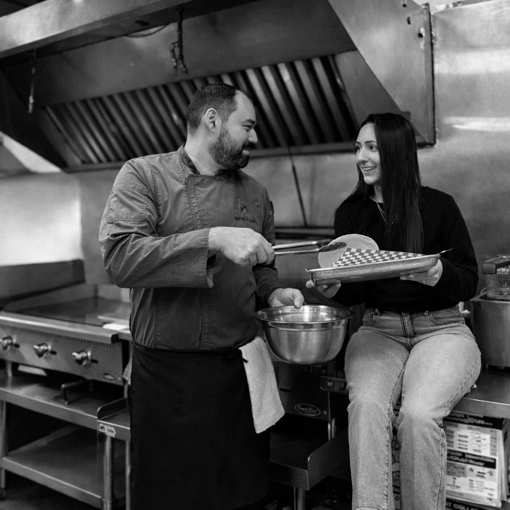 A chef and a woman exchanging smiles in a commercial kitchen, with kitchen equipment and a hood ventilation system overhead.