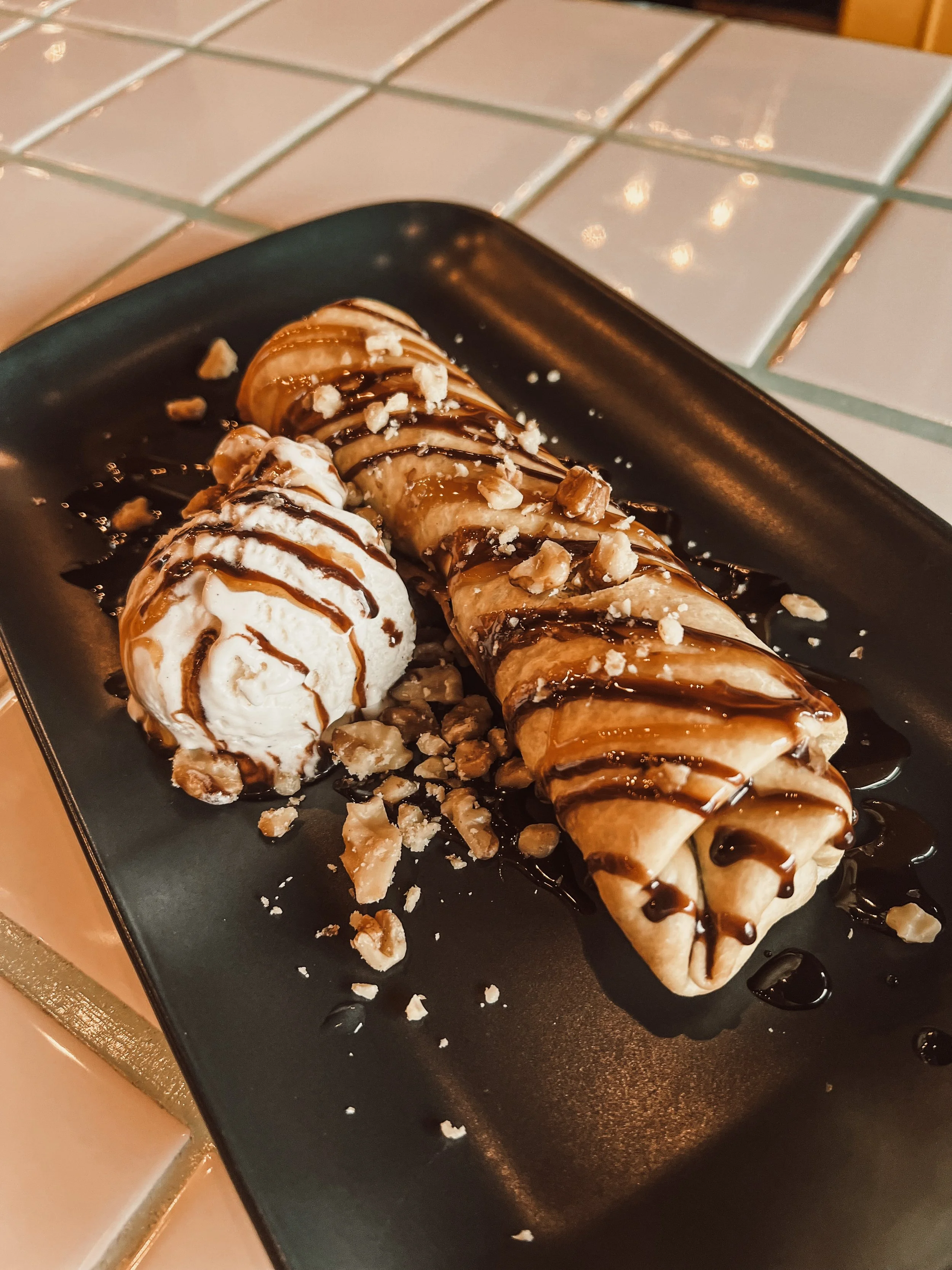 A dessert plate with a nut-filled banana spring roll drizzled with chocolate sauce and a scoop of vanilla ice cream topped with chocolate syrup, garnished with chopped nuts.