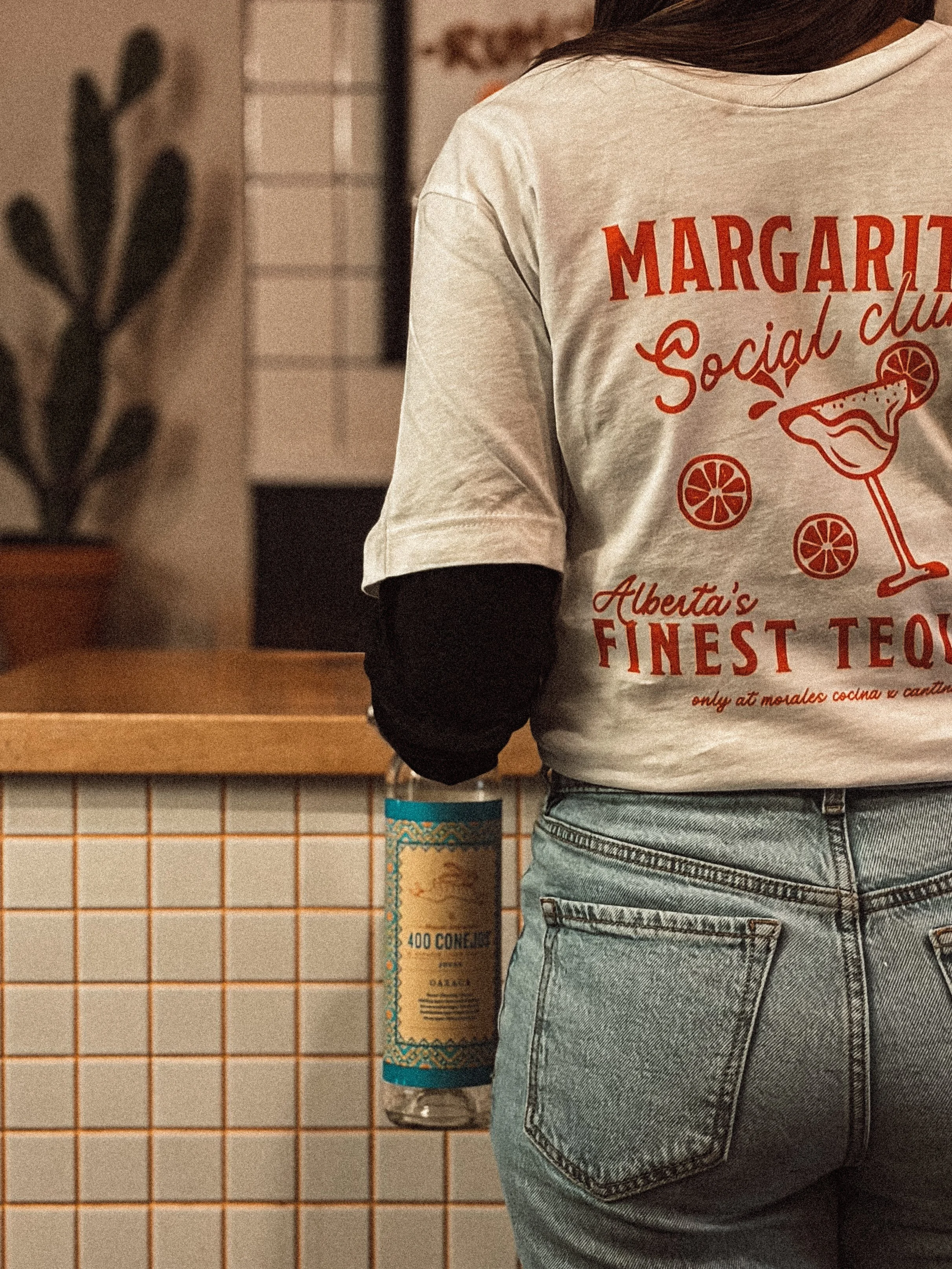 A person wearing a gray t-shirt with red text and graphics, standing at a counter in a cafe or restaurant. The t-shirt advertises a social club and tequila brand. The person is also wearing light blue jeans, and there is a water bottle hanging from their pocket.