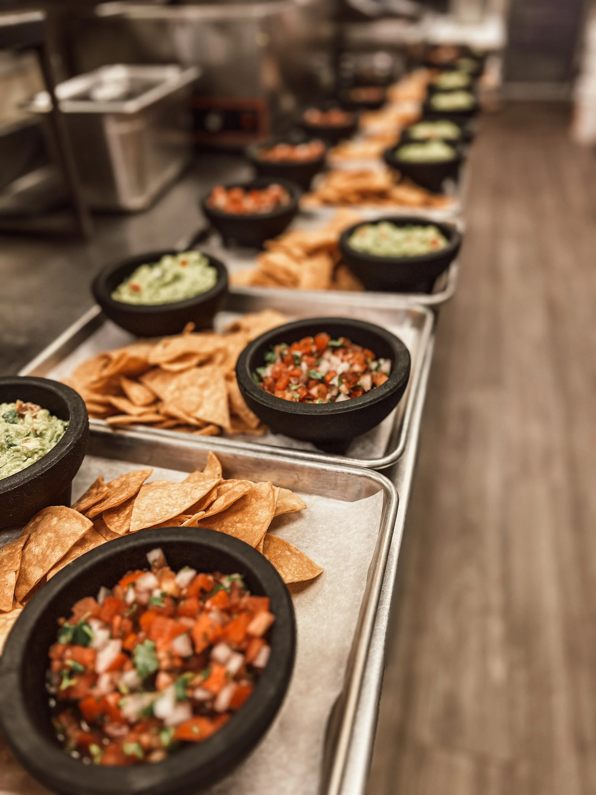 Trays with bowls of guacamole, salsa, tortilla chips, and other Mexican food items in a restaurant kitchen.