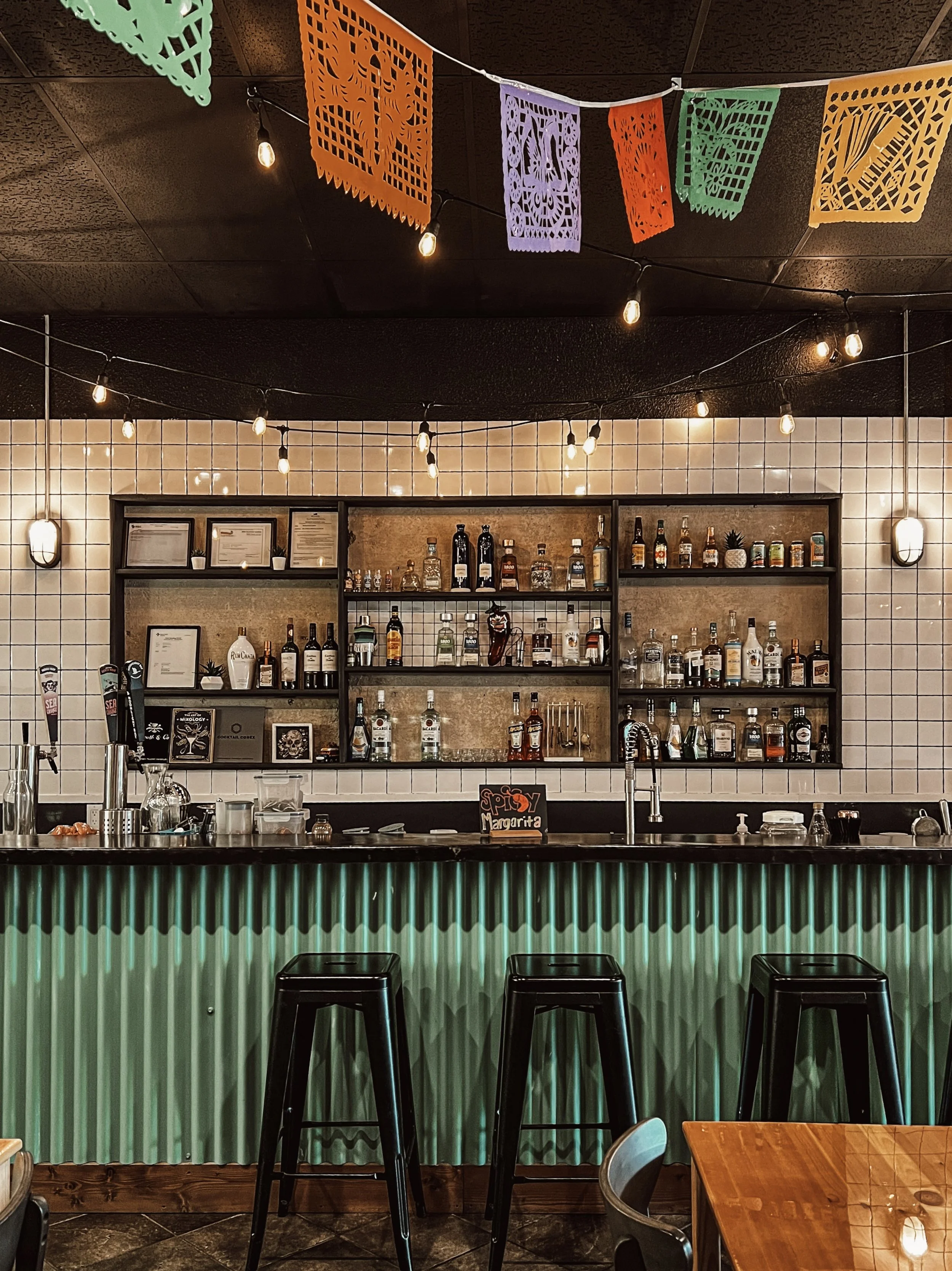 Interior of a bar with a illuminated green corrugated metal front, black bar stools, a tablet on the bar, hanging string lights, decorative Papel Picado banners on the ceiling, shelves with alcohol bottles, a sink, and a chalkboard sign that says 'Spicy Margarita'.