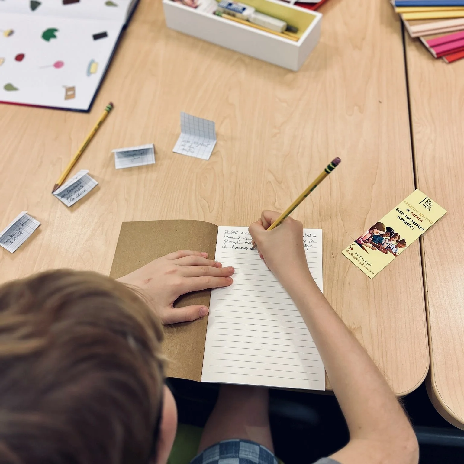 Child writing in a notebook at a wooden table with various art supplies, including colored paper, stickers, and handwritten notes.
