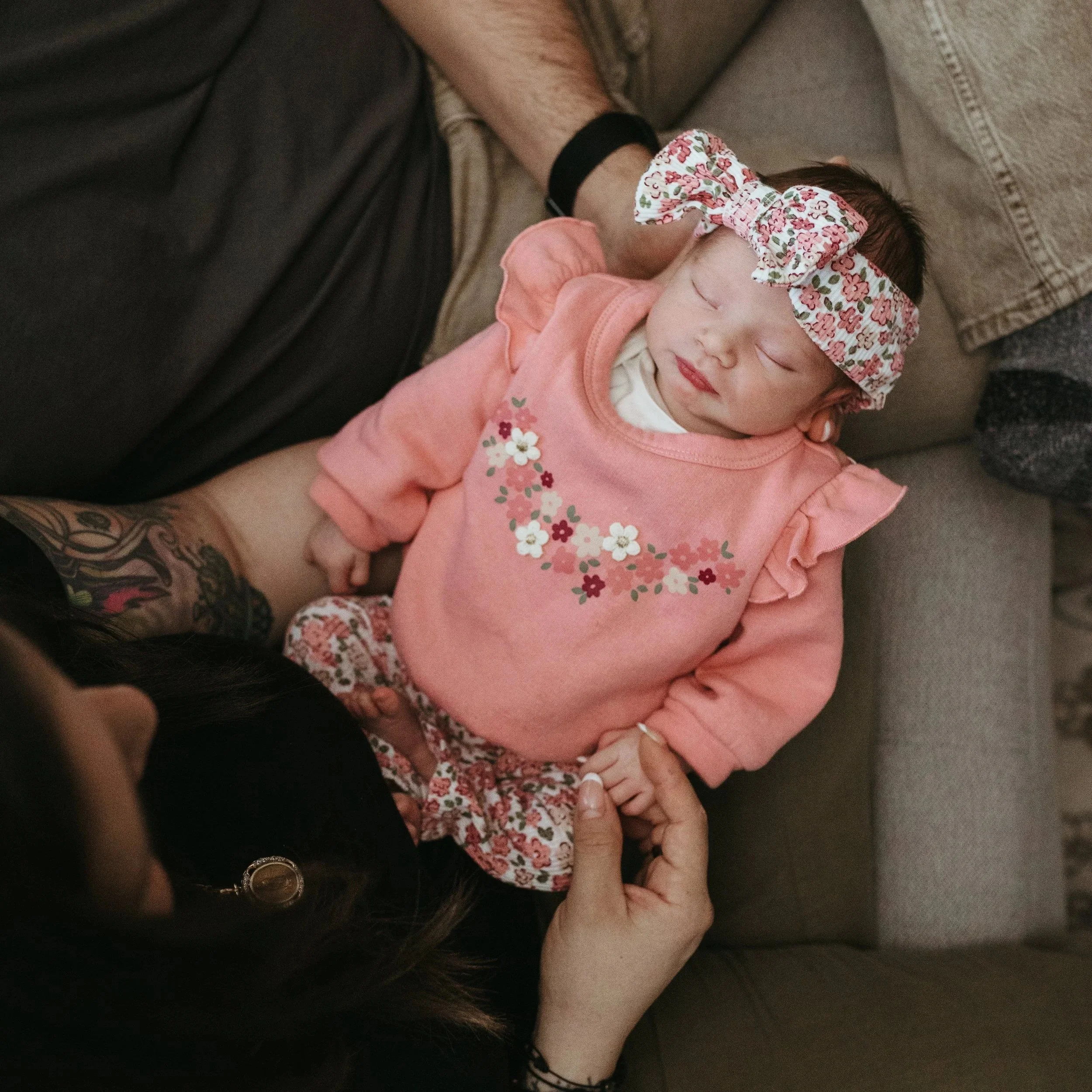 A newborn baby girl with long brown hair wearing a floral headband and a pink shirt with floral embroidery is peacefully sleeping with her head resting on an adult's arm. The adult is holding the child's head gently while lying on a brown couch.