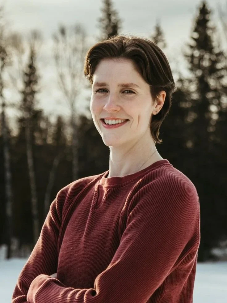 A person with short brown hair smiling outdoors in a wintry landscape.