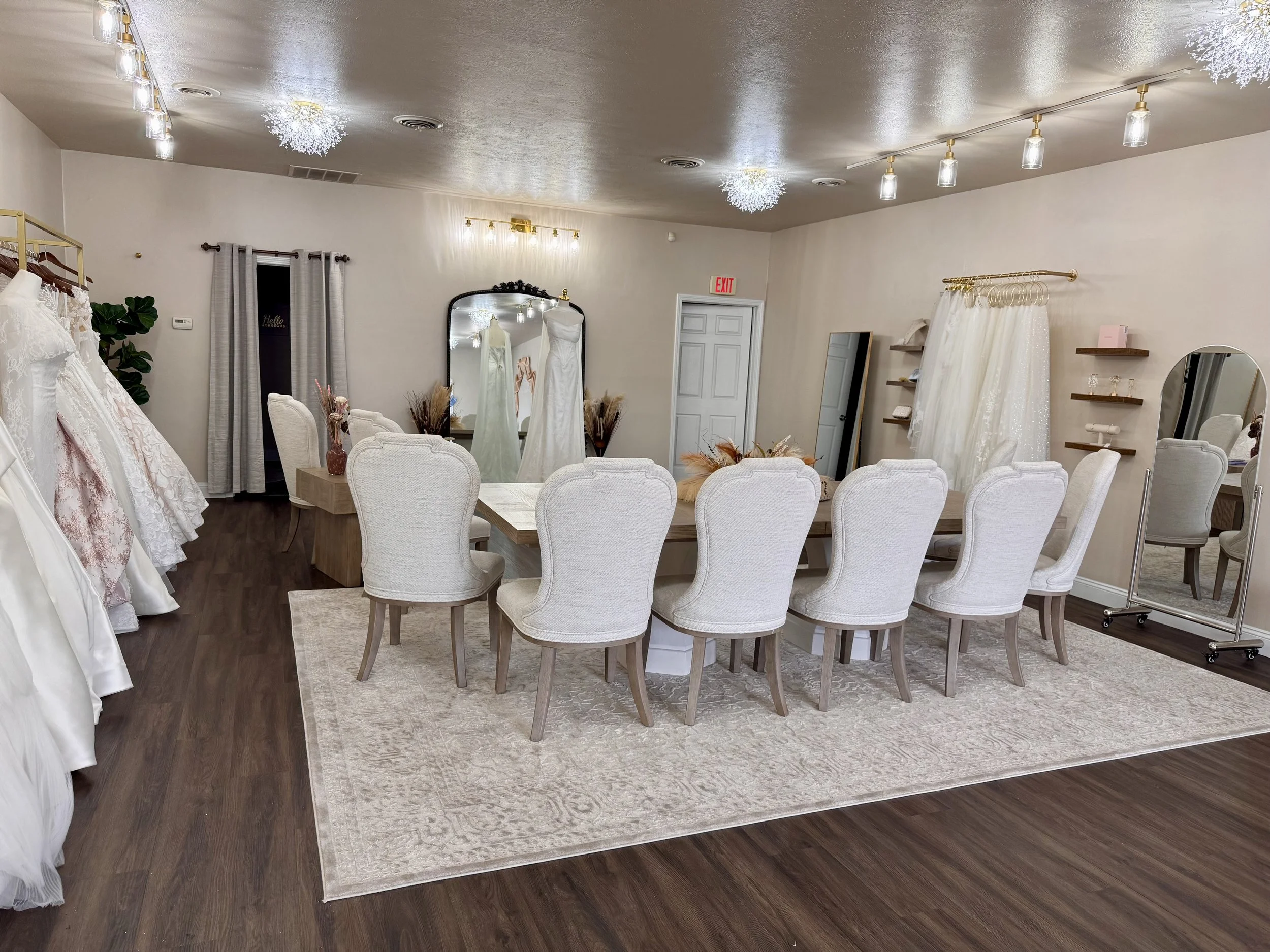 Indoor space decorated with wedding dresses on mannequins near the window, with chairs and a table with flowers, and soft lighting from ceiling fixtures and decorative lights.