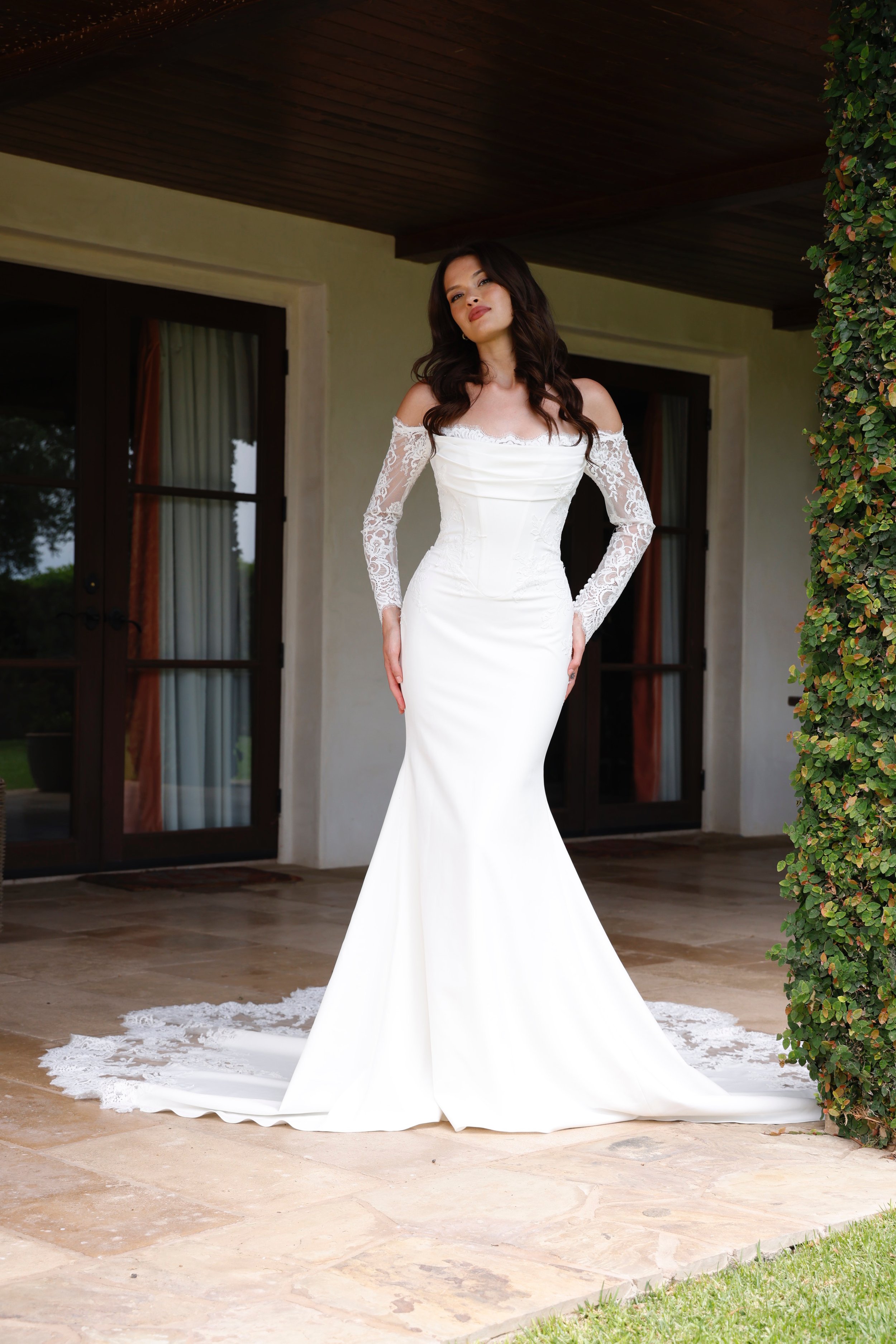 Woman in a white wedding dress standing outside on a patio area with a wood ceiling, next to a green bush, near a building with glass doors and beige walls.