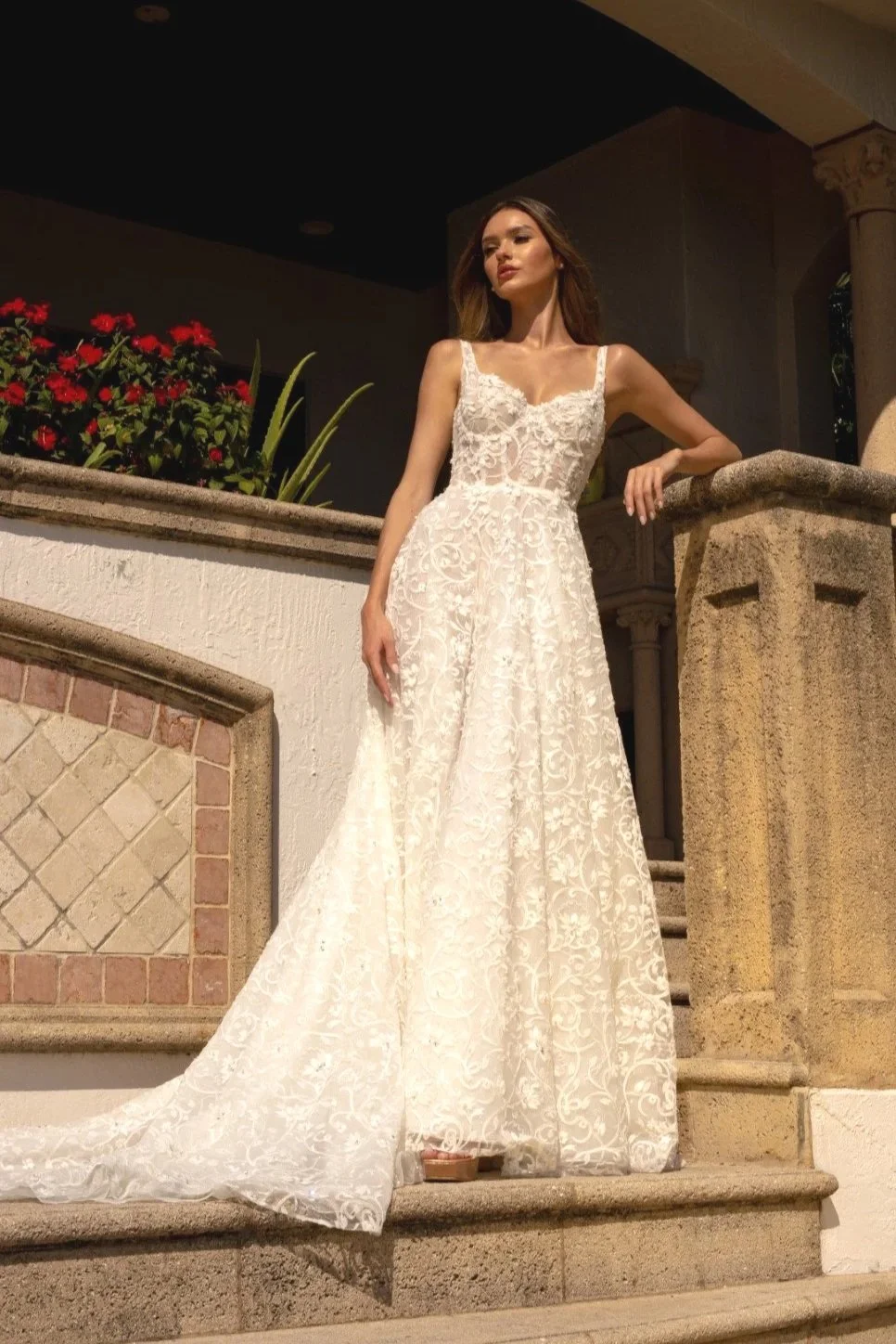 A woman wearing a white lace wedding dress standing on stone steps outside a building with columns and red flowers.