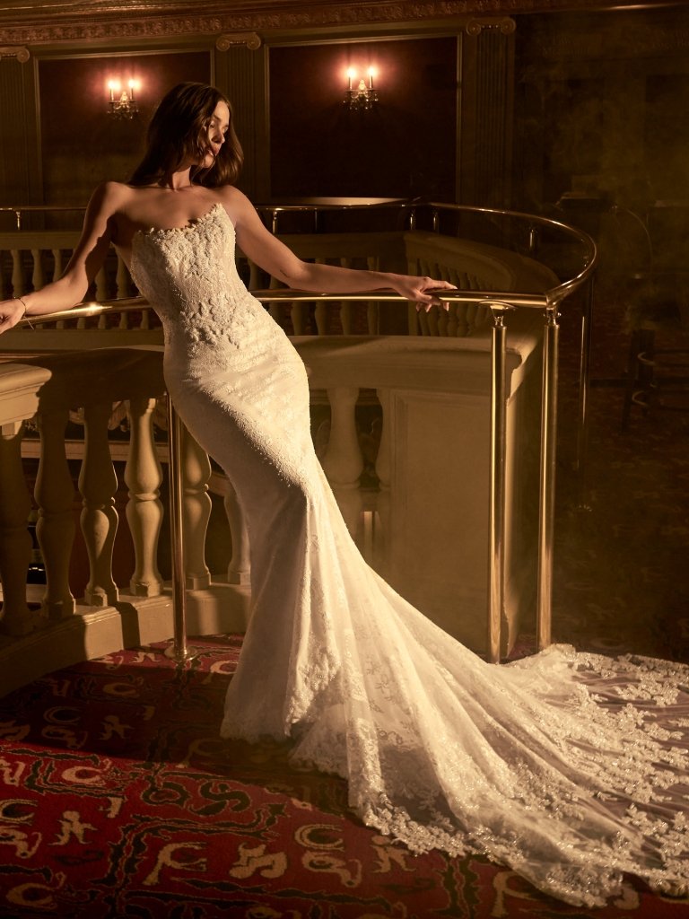 Bride in white wedding dress with train, standing by large window with brick wall background, holding sheer veil.