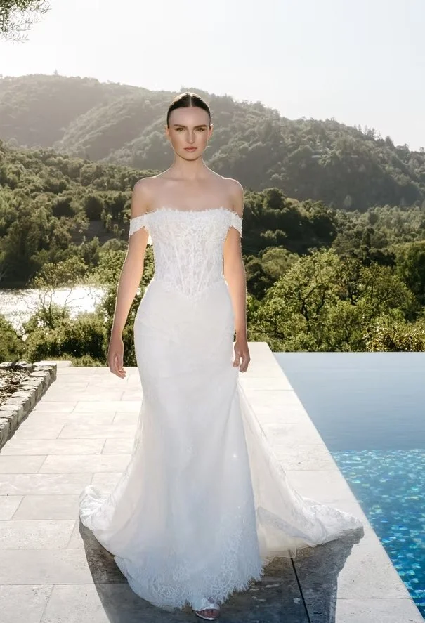 A woman in a strapless white wedding dress standing outdoors by a swimming pool with a mountainous landscape in the background.