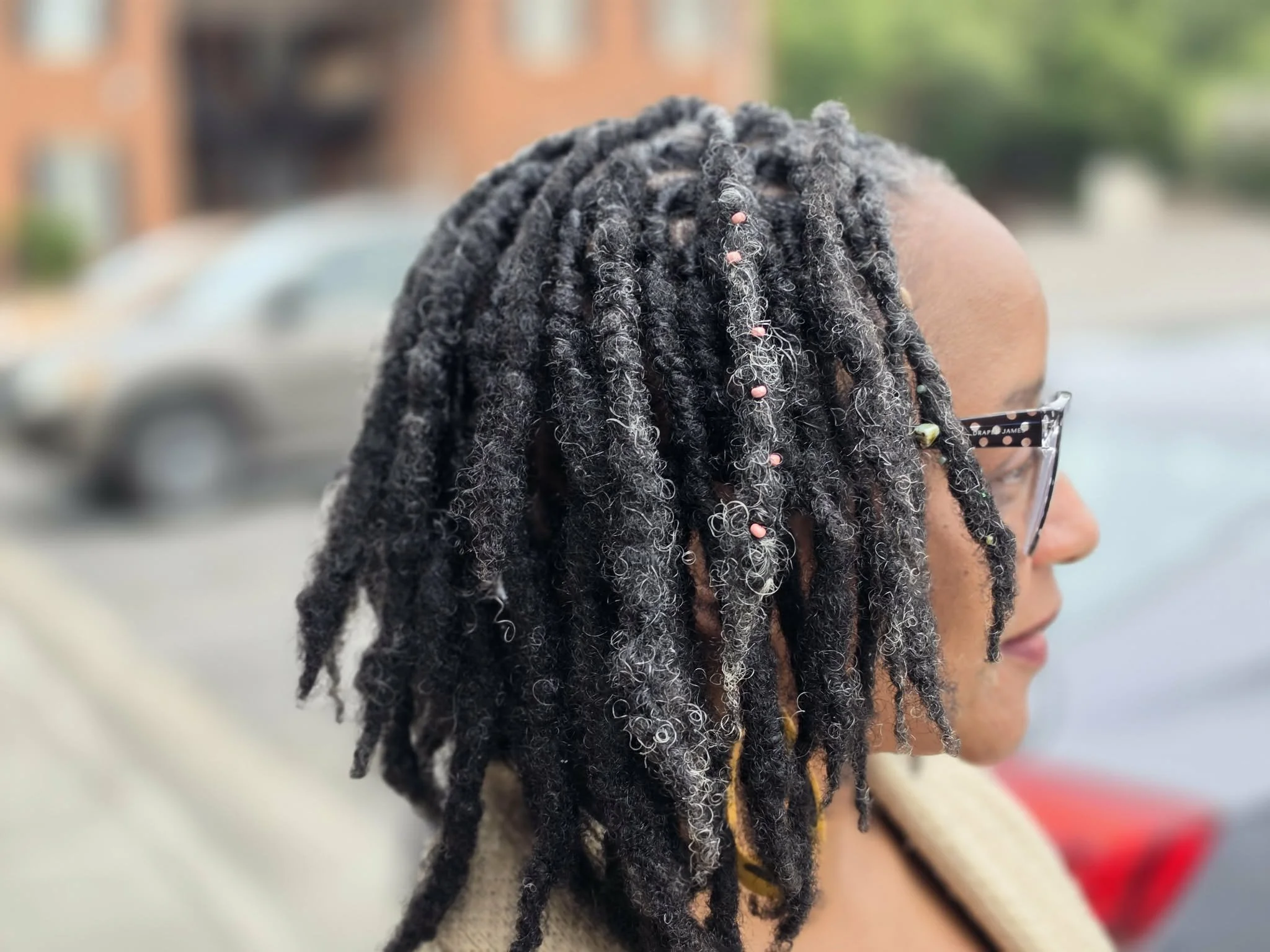 Side profile of a woman with naturally styled dreadlocks with Hair Sprinkles loc adornments, wearing glasses, outdoors with blurred background.