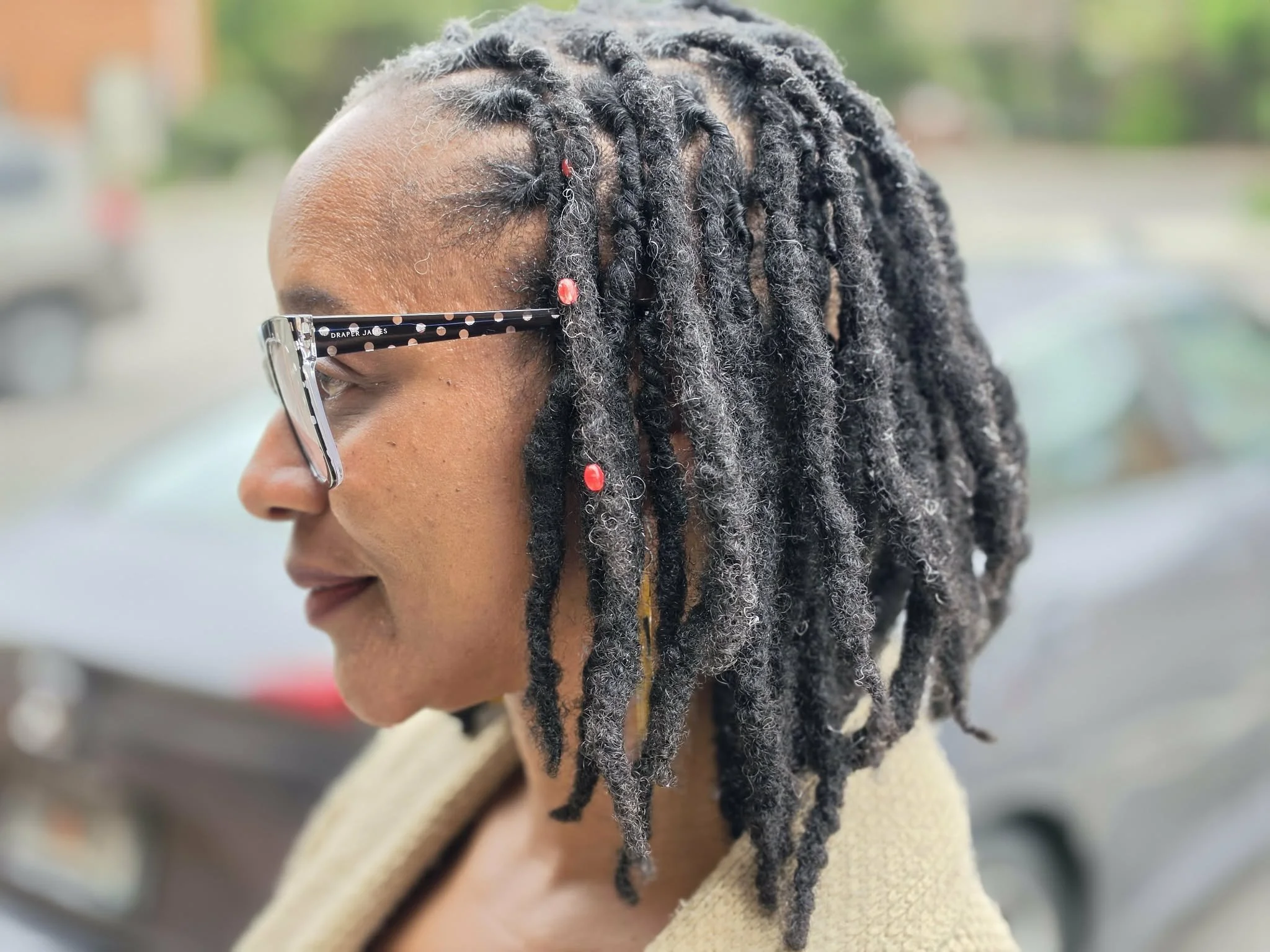 Side profile of a woman with dreadlocks with Hair Sprinkles loc adornments, wearing black and white glasses and a beige coat outdoors.