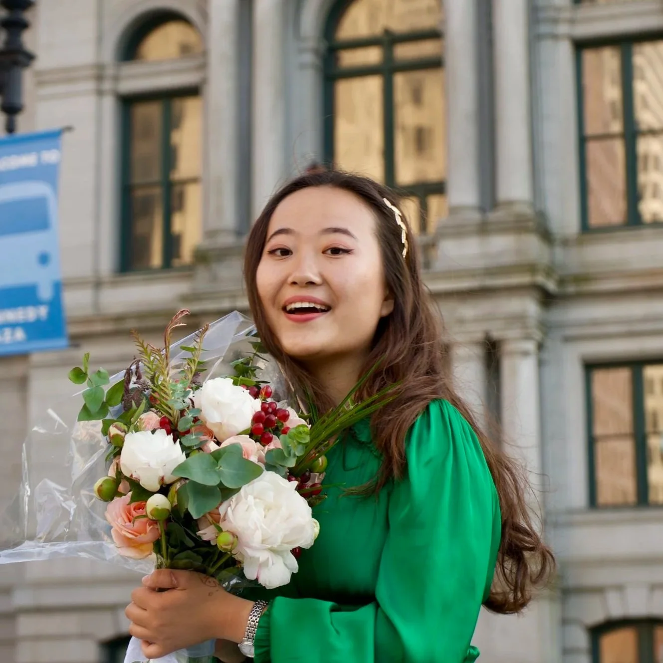 A young woman in a green dress holding a bouquet of flowers outdoors in front of a historic building.