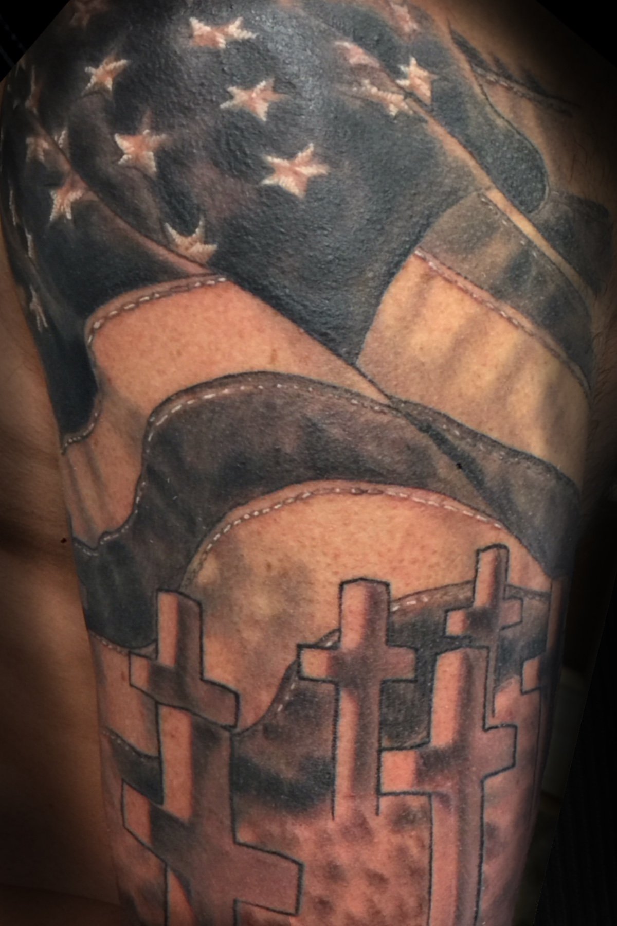 Black and grey American flag and cross tombstone tattoo on an upper arm. Tattoo art by Daniel Stewart at Signature Rose Professional Tattooing in Muncie Indiana.