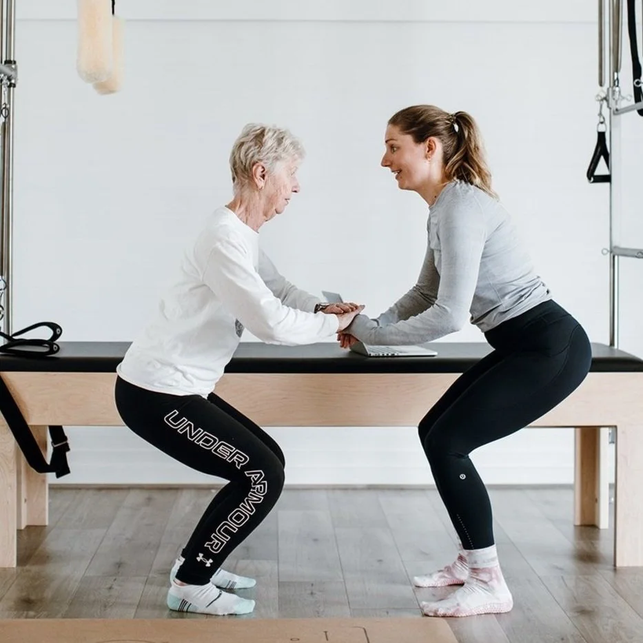 Qualified Pilates instructor instructing an older women in a squat in front of a pilates trapeze table
