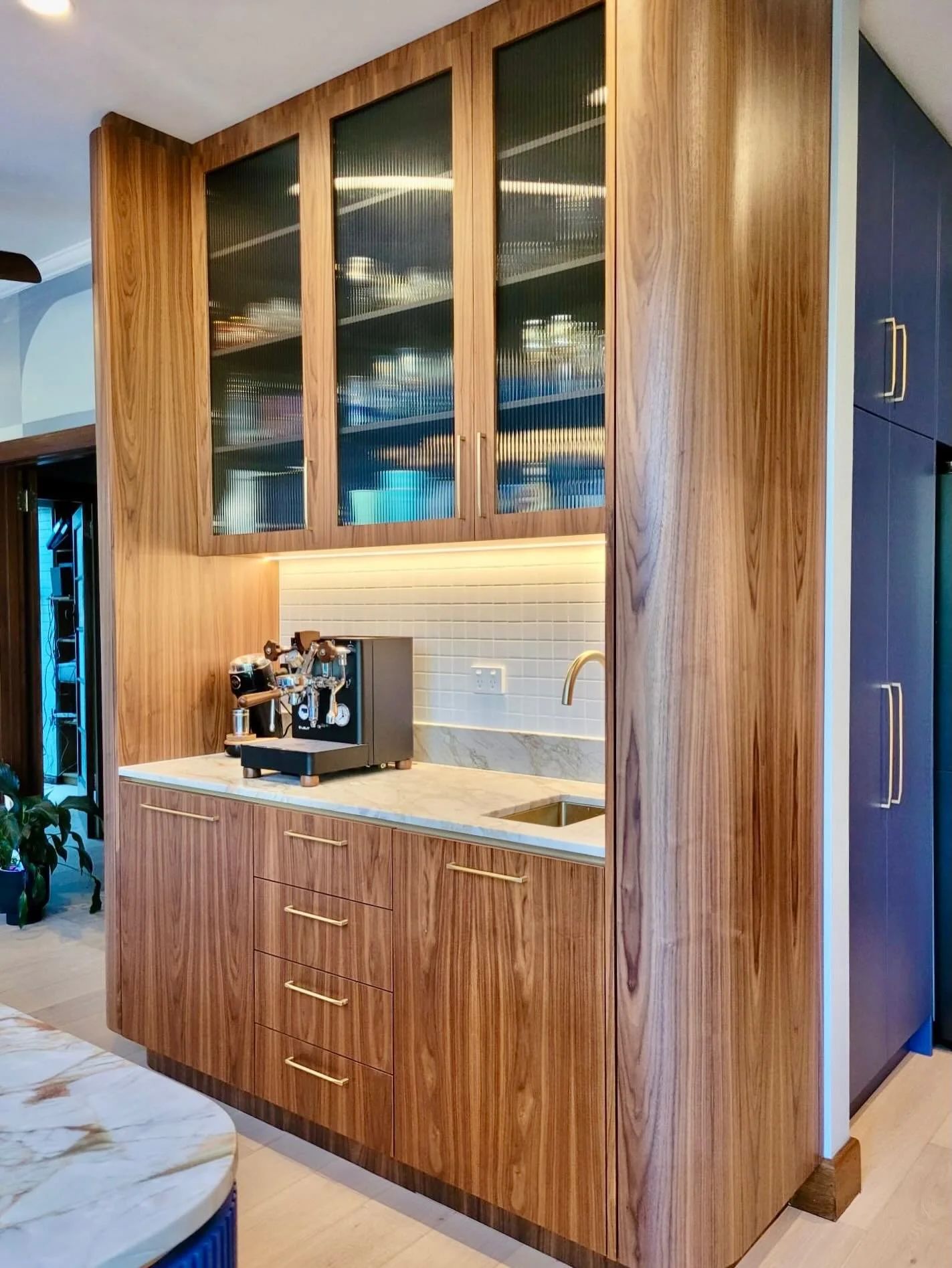 A coffee bar that feels as good as it looks.
Natural walnut veneer wraps the space in warmth, while fluted glass softens the view and adds a gentle, layered glow. Integrated lighting brings a quiet sense of calm to the everyday ritual.
The curved end