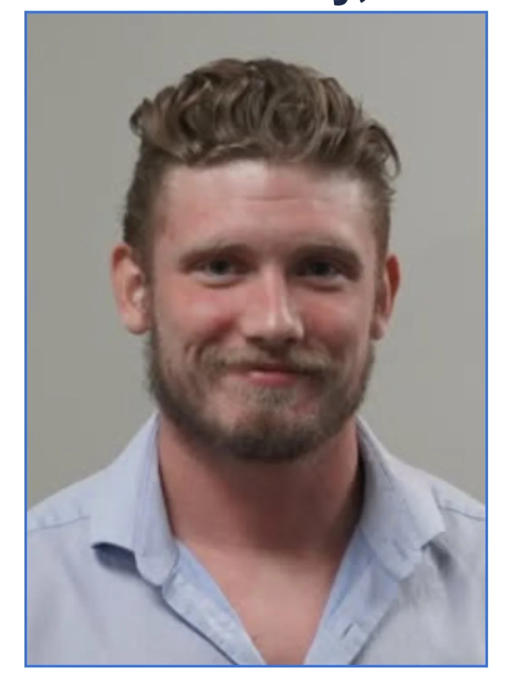 Portrait of a man with short wavy hair, a beard, and a light blue button-down shirt, smiling slightly against a plain background.