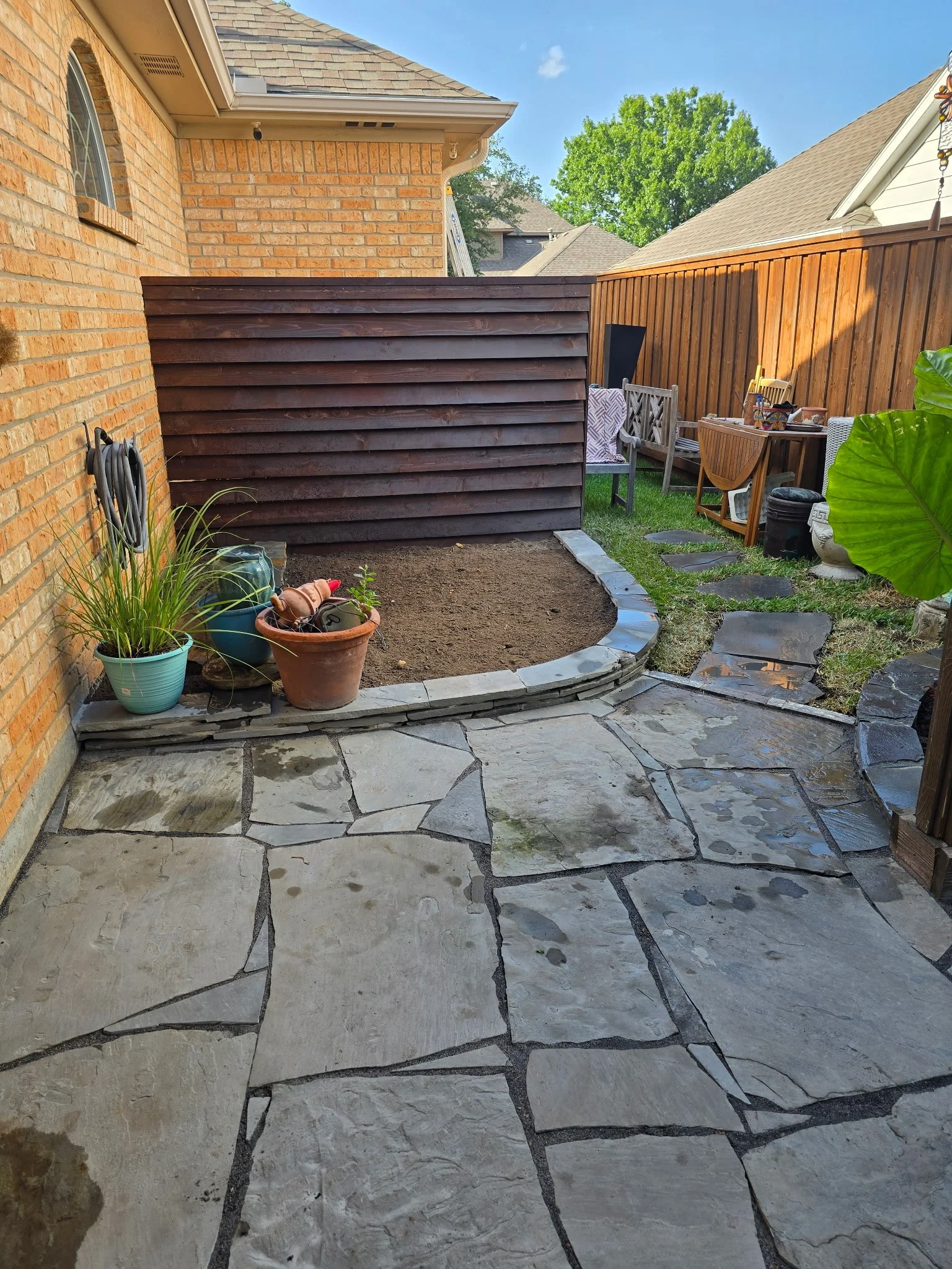 Backyard patio with irregular stone pavers, some soil without grass, and a small garden bed with potted plants. There are outdoor furniture items, a wooden fence, and part of a brick house.