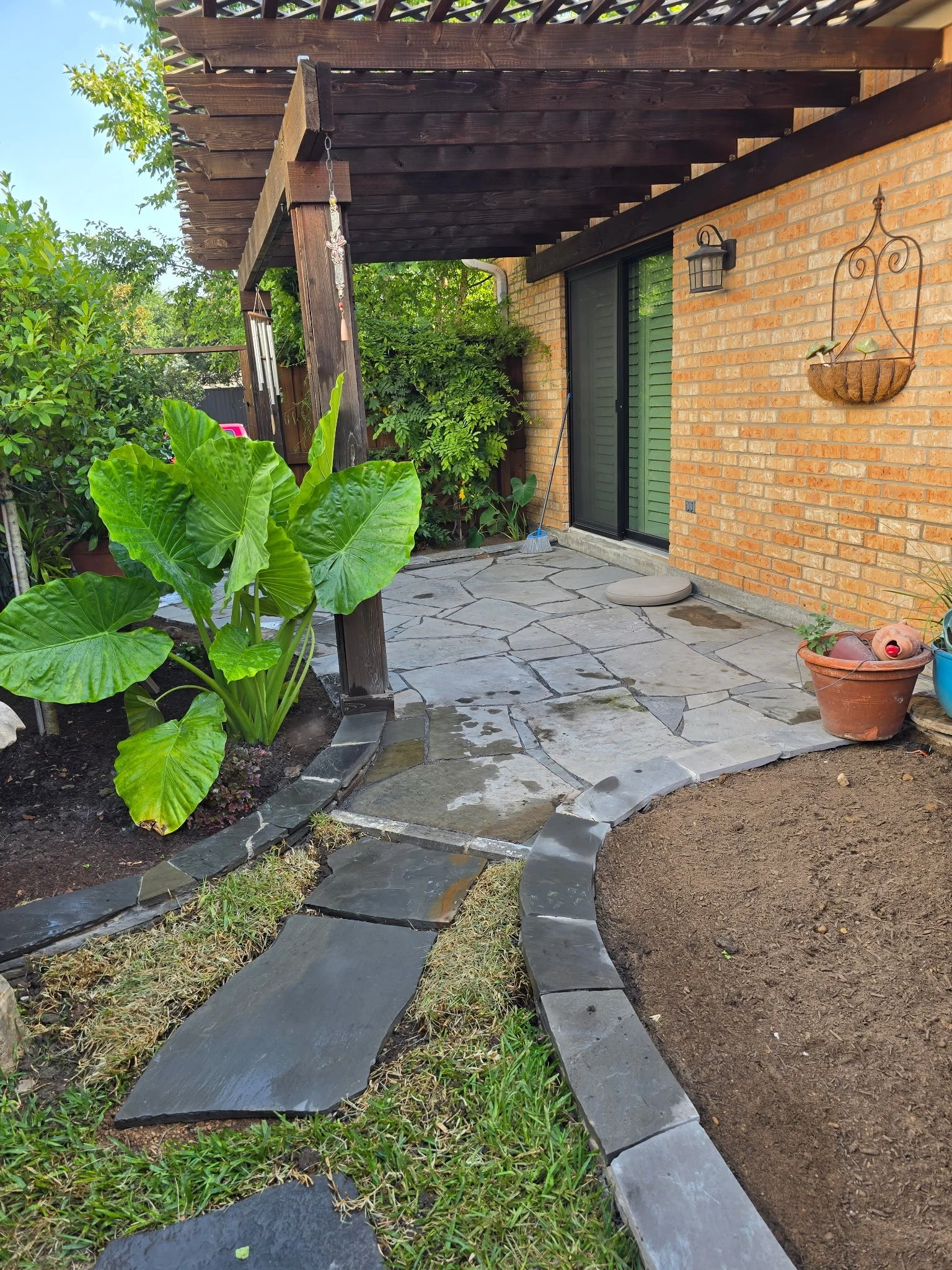 A backyard patio with a stone pathway, potted plants, green shrubs, and a brick house wall with a sliding glass door. There is a wooden pergola overhead, and gardening tools are visible near the door.