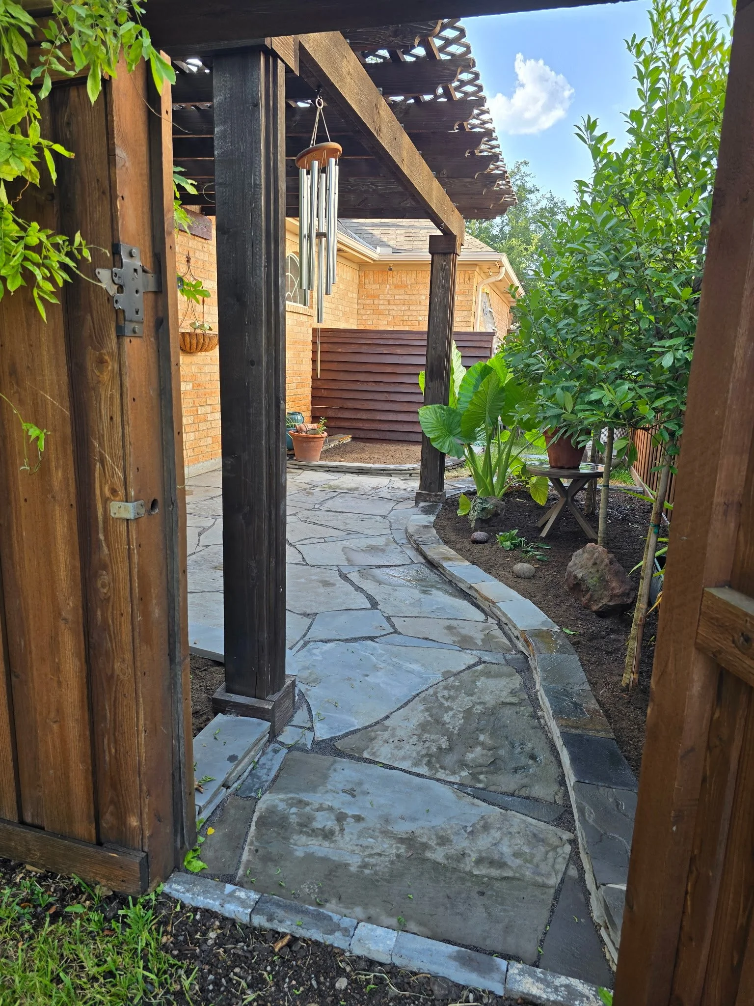 View of a backyard garden with a stone pathway, plants, and a wind chime hanging from a wooden structure.