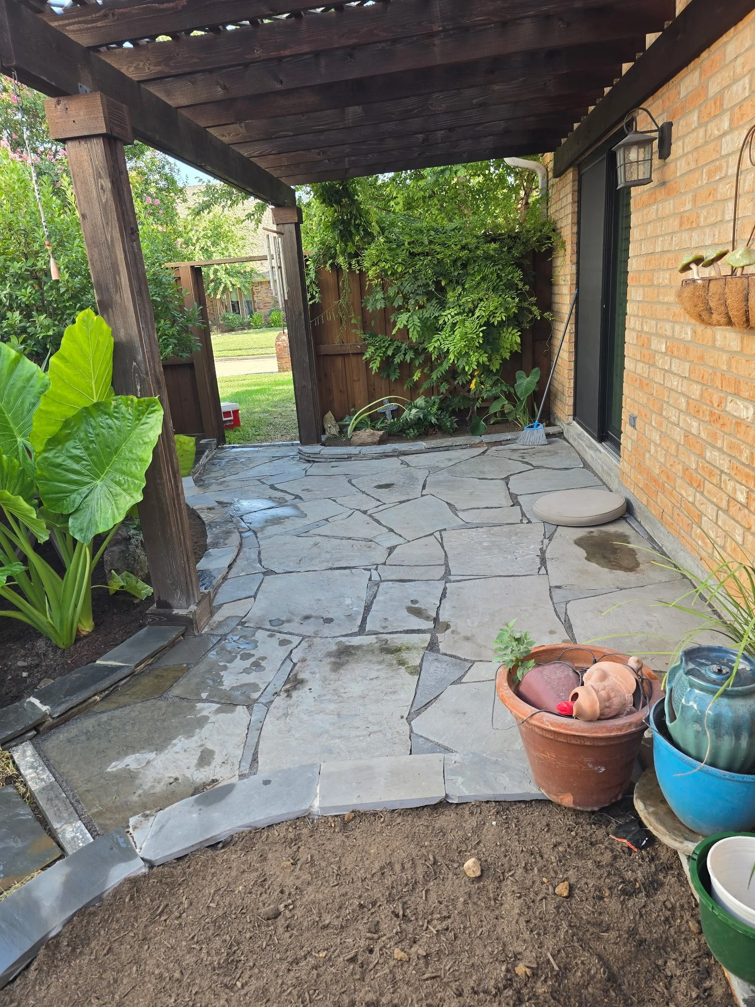 A backyard patio with a stone floor, some plants, a brown wooden fence, and an overhang with dark wood beams. There are gardening pots and a gardening tool leaning against the house wall, which is made of orange brick.