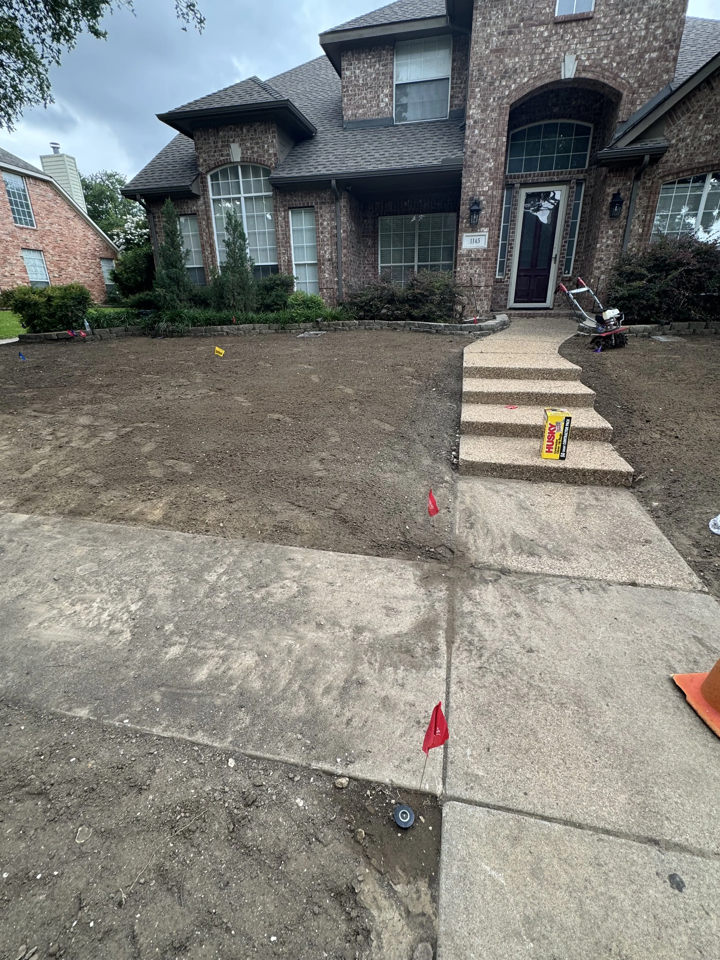 Front yard of a house under landscaping, with freshly dug dirt, a concrete walkway and stairs leading to the front door, and a box of Husky lawn seed on the walkway.