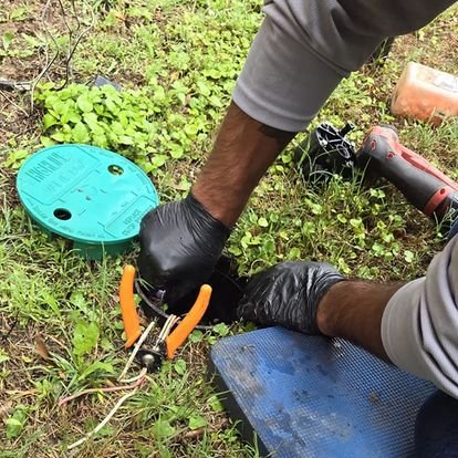 Person using pliers to work on underground wiring or cable in grassy area, wearing black gloves and gray sleeves.