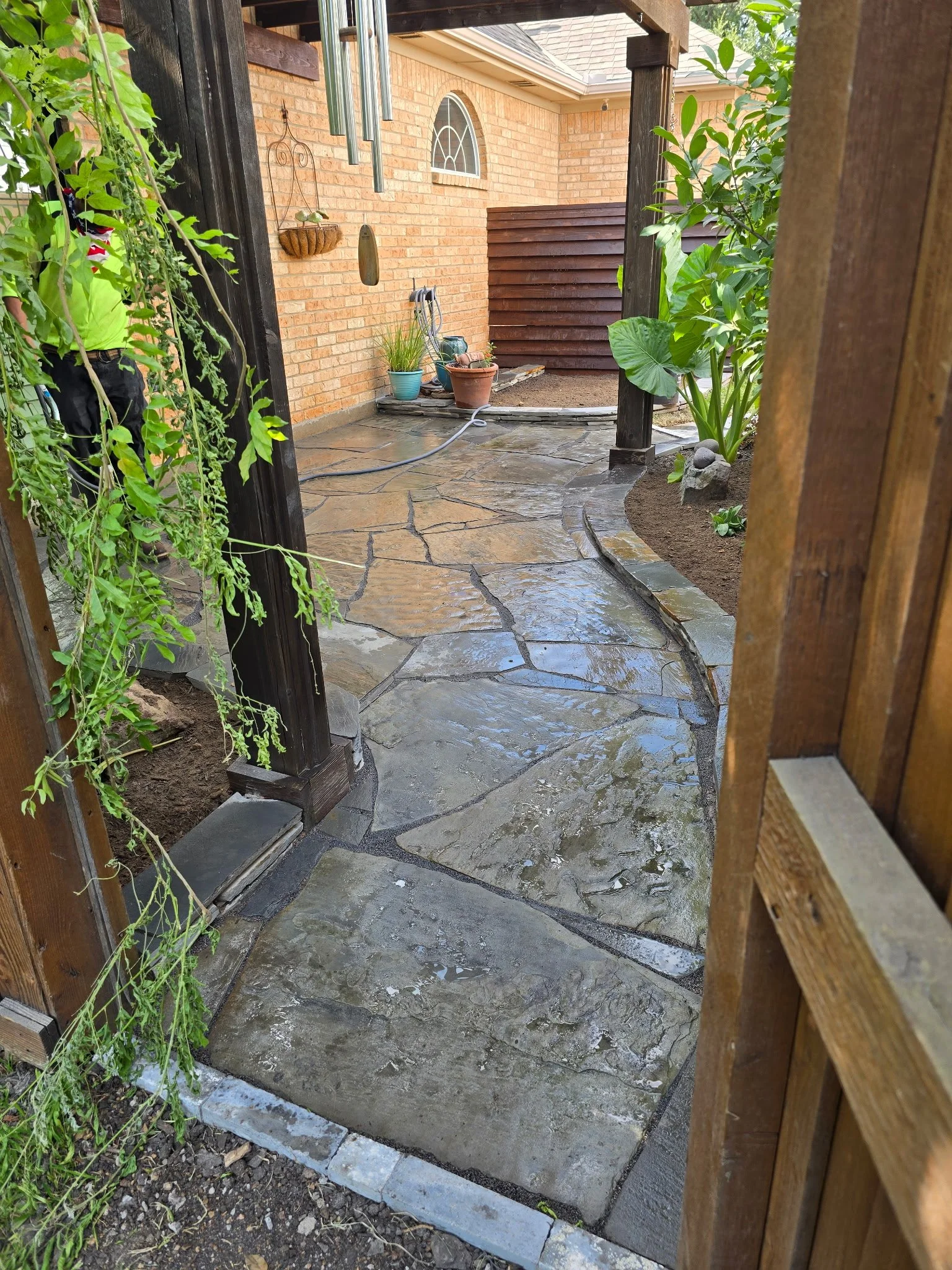 View of a backyard with a stone pathway, potted plants, a hanging basket, wind chimes, and a wooden fence, with a brick house wall in the background.