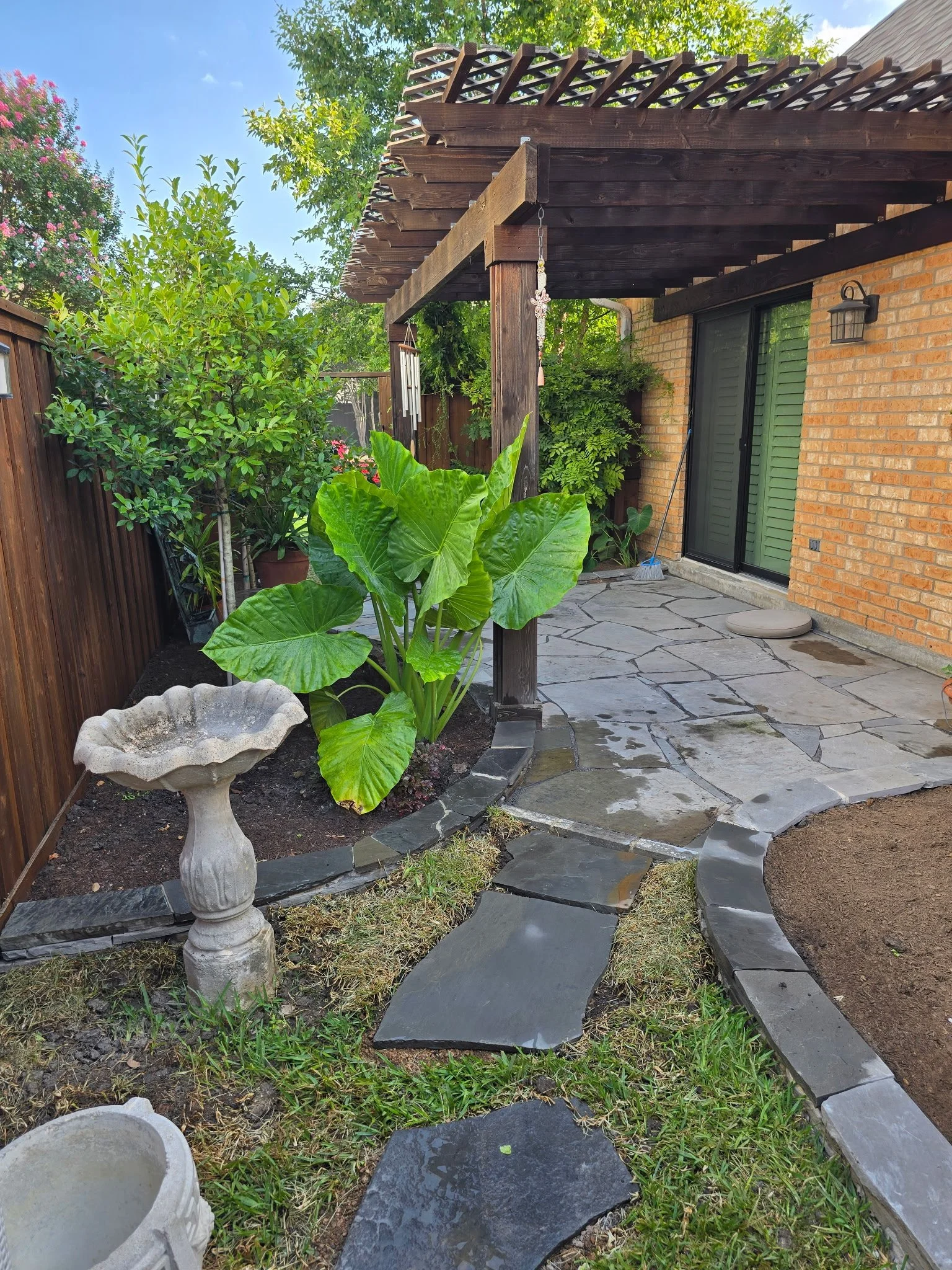 A backyard patio with flagstone flooring, a brick house wall with a sliding glass door, a wooden pergola, green leafy plants, a birdbath, and a garden hose.