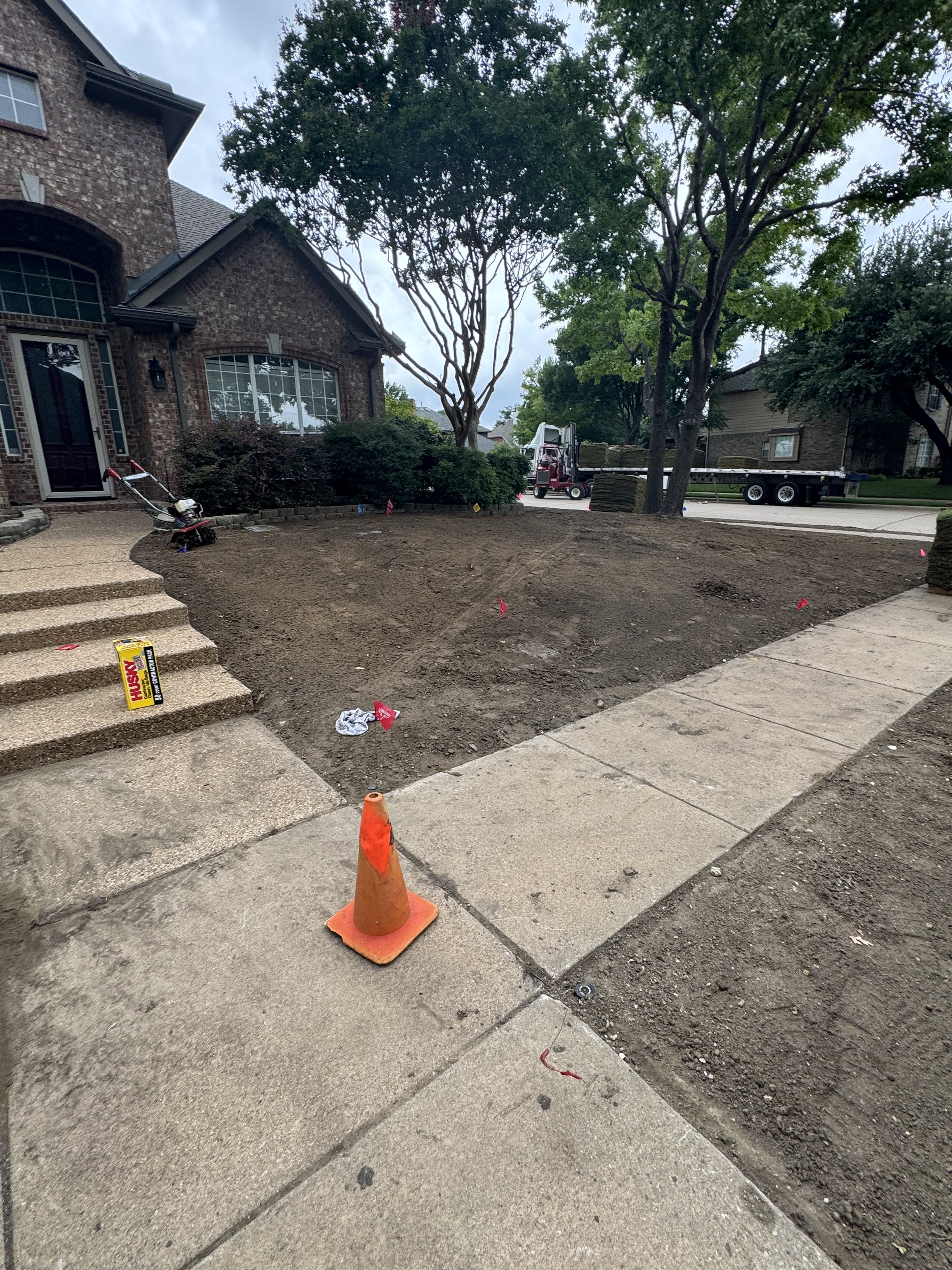 A residential front yard with a garden bed being prepared, orange traffic cone, a box of Husky brand nails, and construction equipment including a flatbed truck and a piece of heavy machinery in the background.