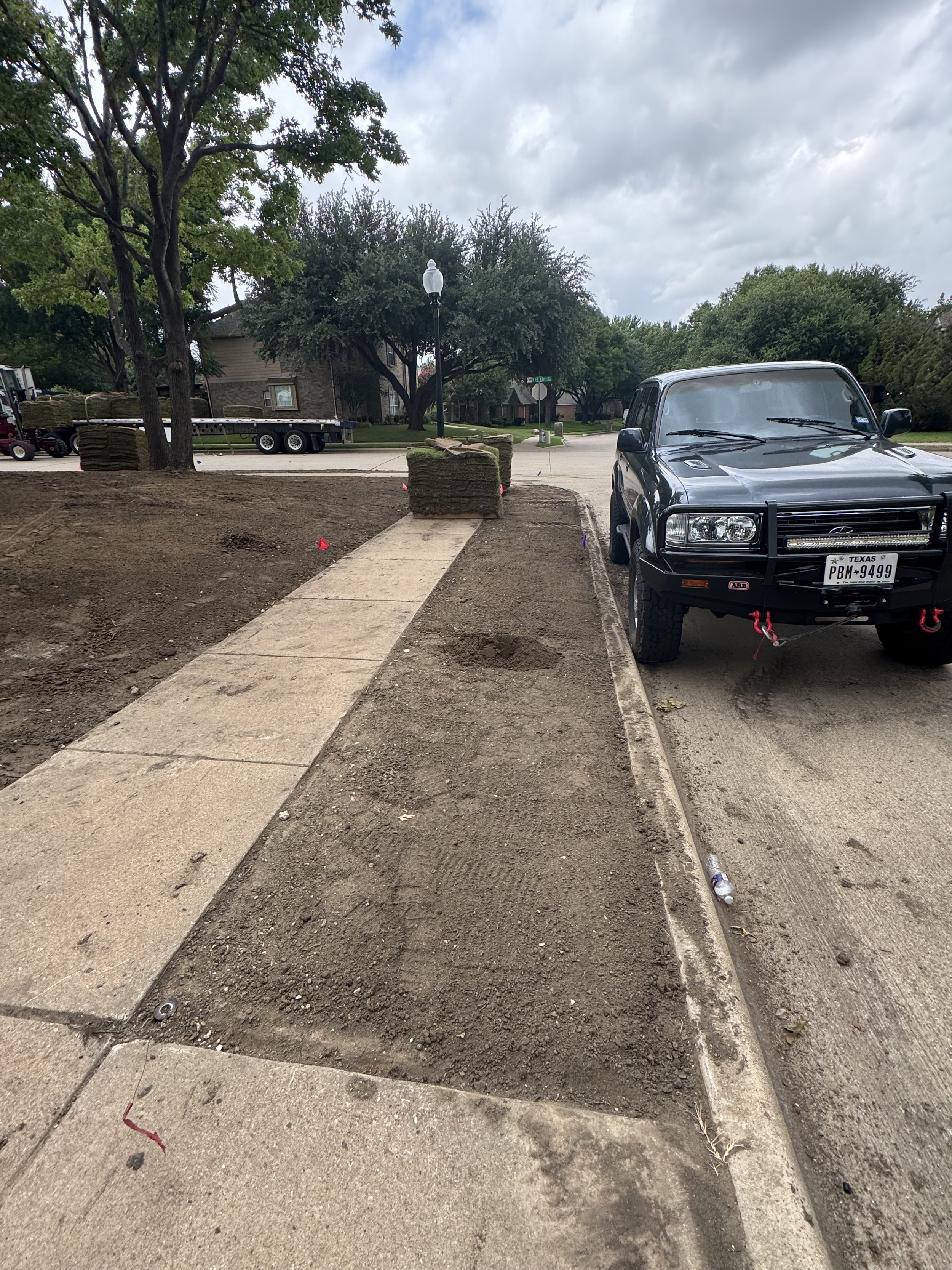 View of a sidewalk and a dirt area with some construction or landscaping materials, a truck with sod rolls in the background, and a black SUV parked by the curb. Trees and residential houses are visible under a cloudy sky.