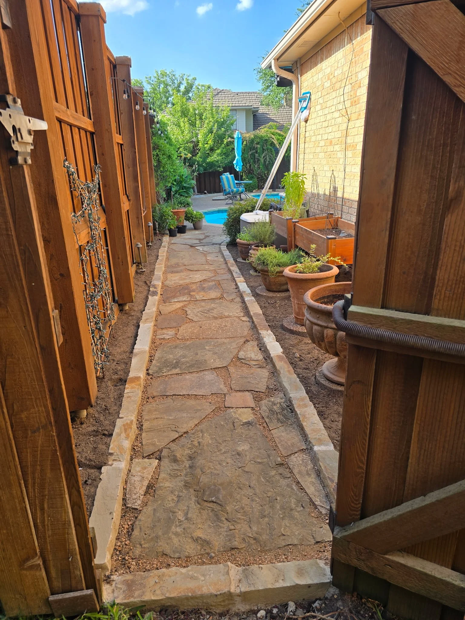 A stone pathway leading to a backyard with plants, potted flowers, trees, and a swimming pool with a blue umbrella and patio chairs, enclosed by a wooden fence and brick house on right.