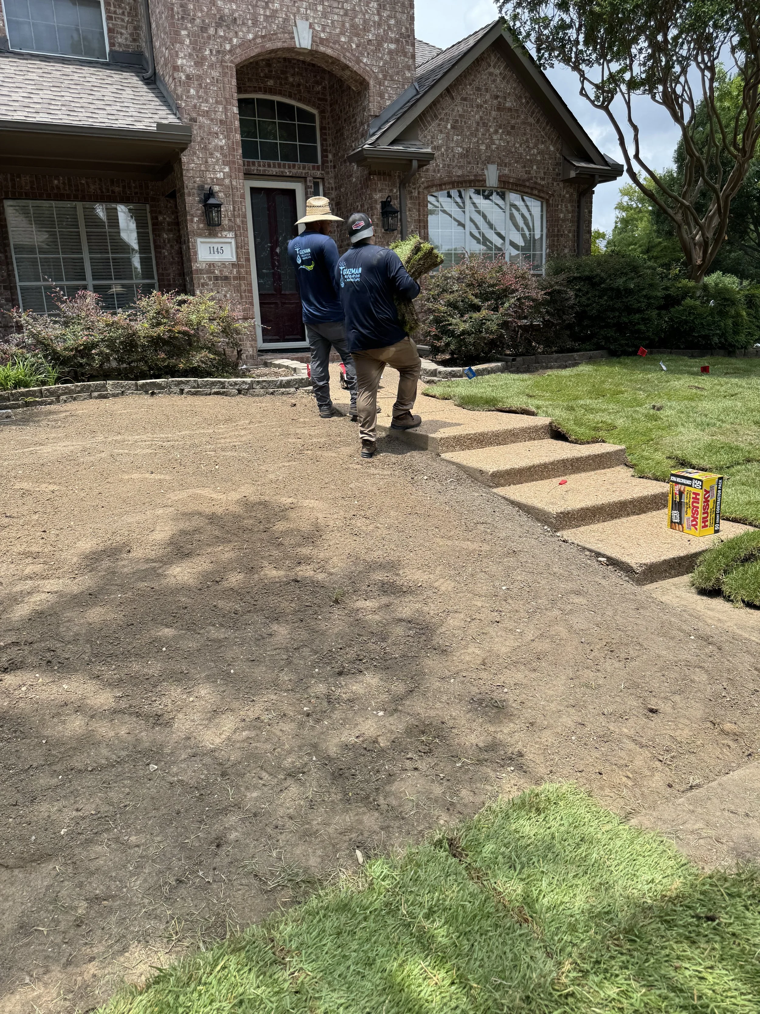 Two workers gardening in front of a brick house, laying sod on the yard, with some gardening supplies on the ground.