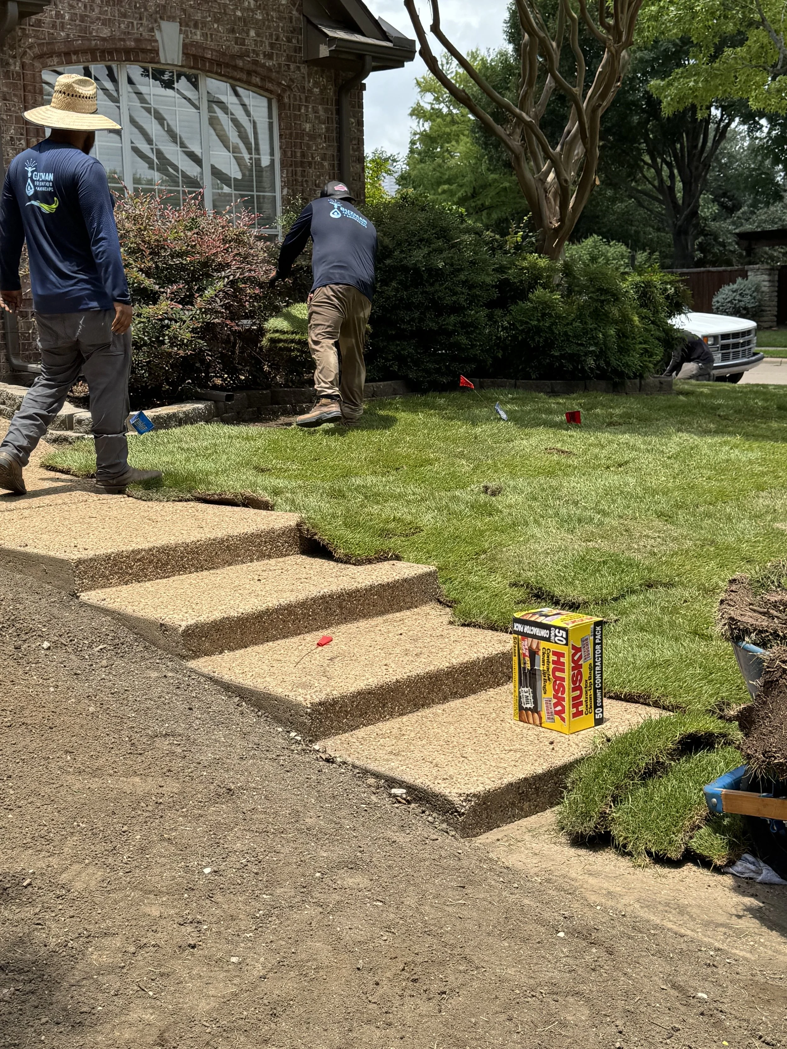 Three men working on landscaping in front of a brick house, installing grass sod on the yard, with steps leading up to the house and plants along the house wall. One man is wearing a straw hat, another is bending over near plants, and the third is kneeling near a white truck. There is a box of grass sod on the ground.