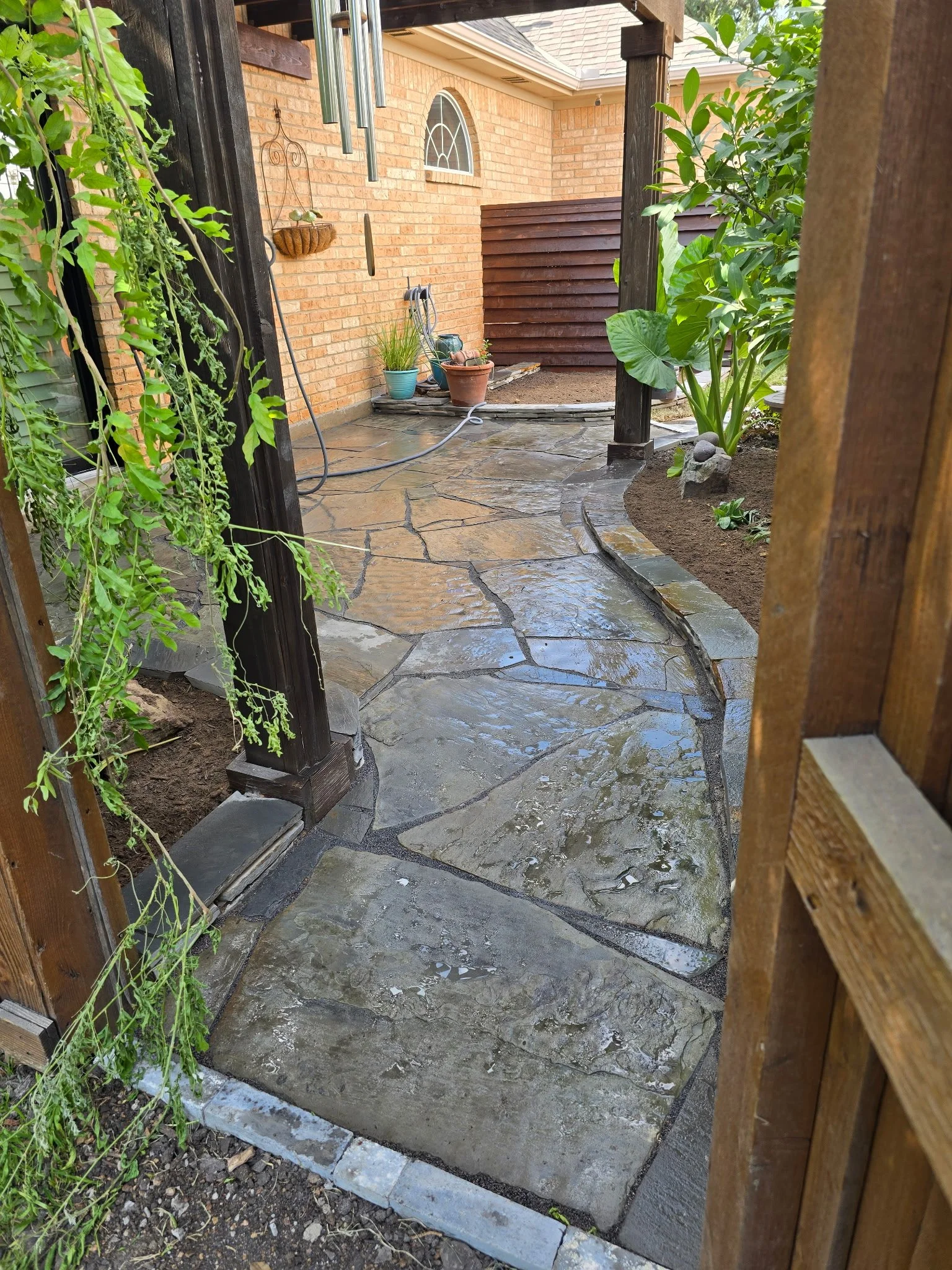 View of a small patio area with a stone pathway leading to a brick house. The patio has a water hose, potted plants, and a wind chime hanging from the house wall. The scene is framed by a wooden gate and some green plants.