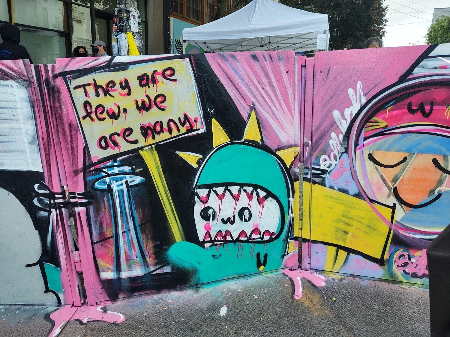 Painting features a cartoonish character holding a sign that reads, "They are few. We are many" near the space needle.