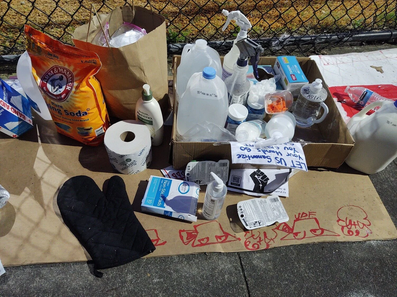 Water, band-aids, baking soda, tissue, and oven mitt are among supplies gathered on a piece of cardboard on a sidewalk.