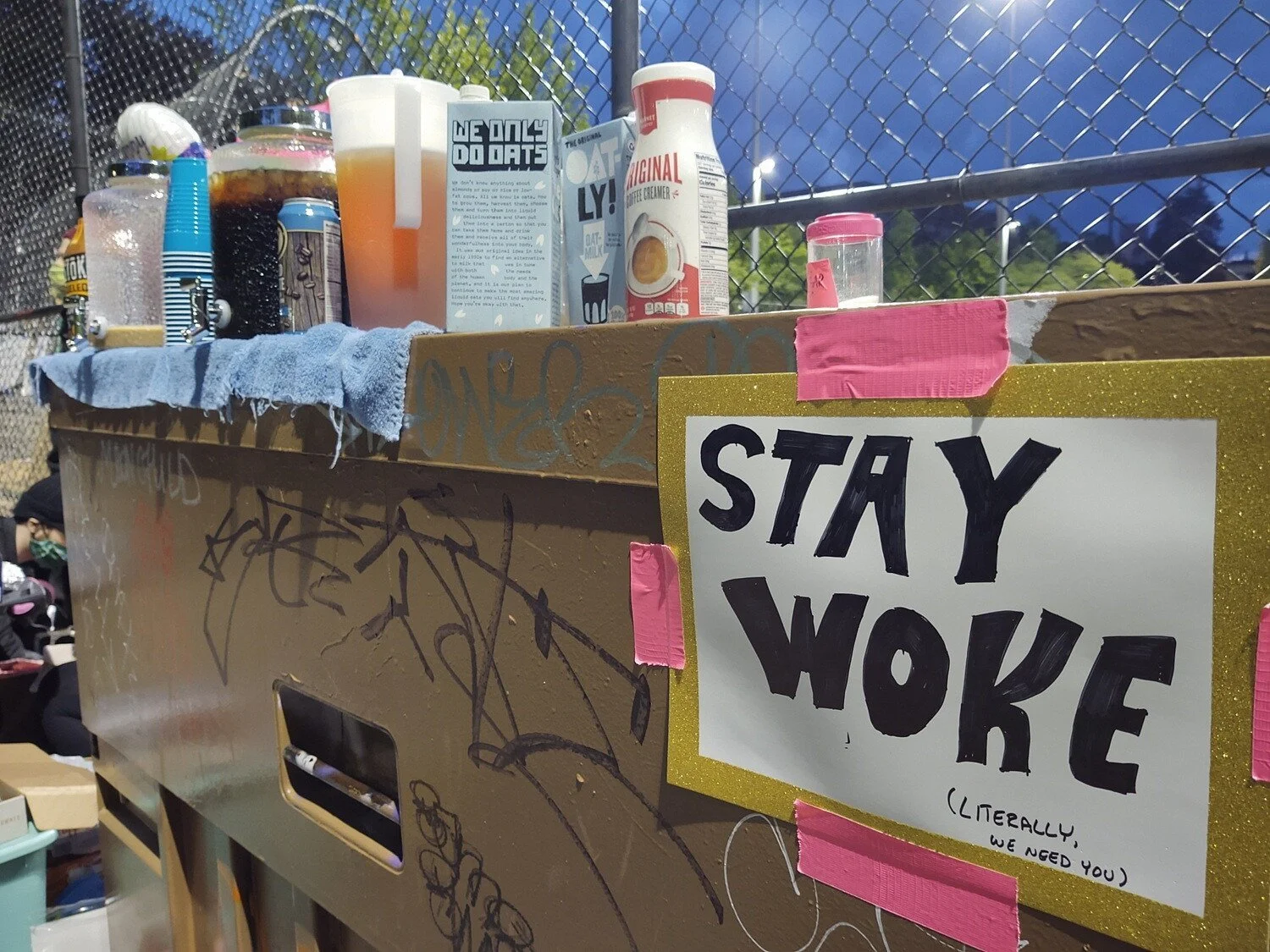 Iced coffee, oat milk, creamer, and cups sit near a hand-made sign reading "Stay Woke (Literally, we need you)”.