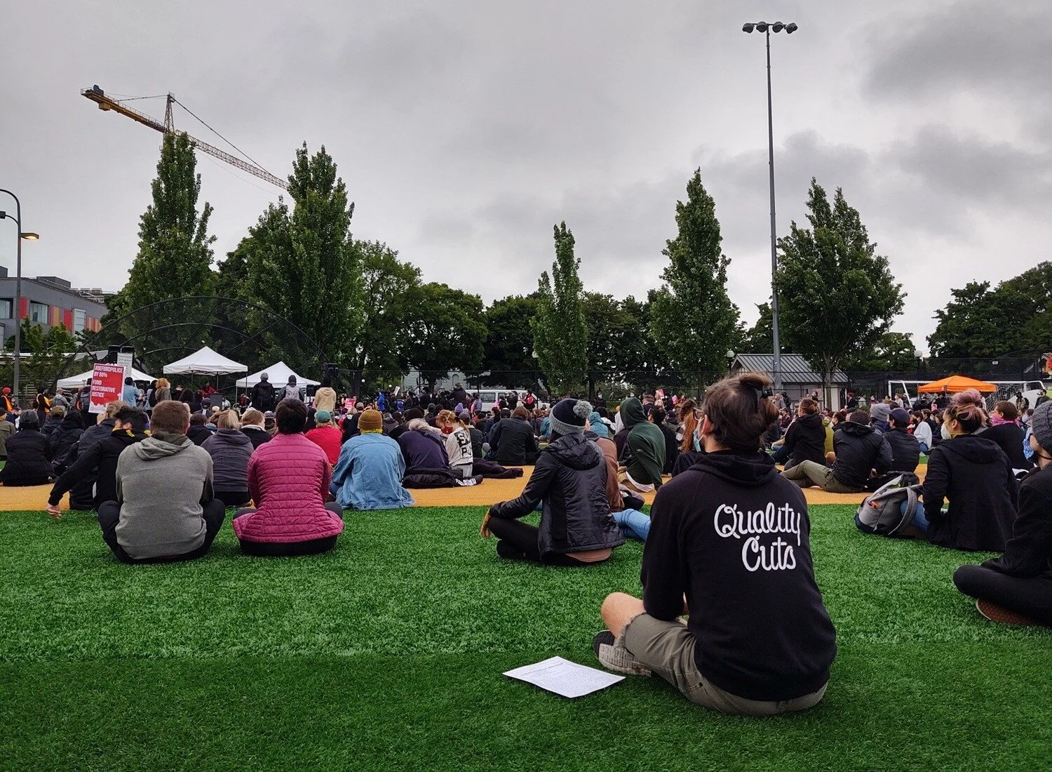 Hundreds to thousands of people sit on a baseball field looking towards white event tents, where speakers are speaking.