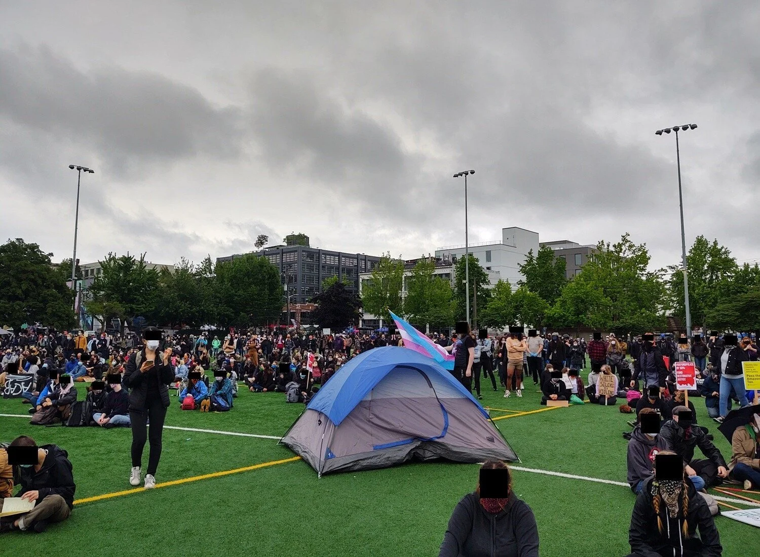 People stand and sit on green astro-turf. A blue tent and a person holding a trans flag are in the center of the frame.