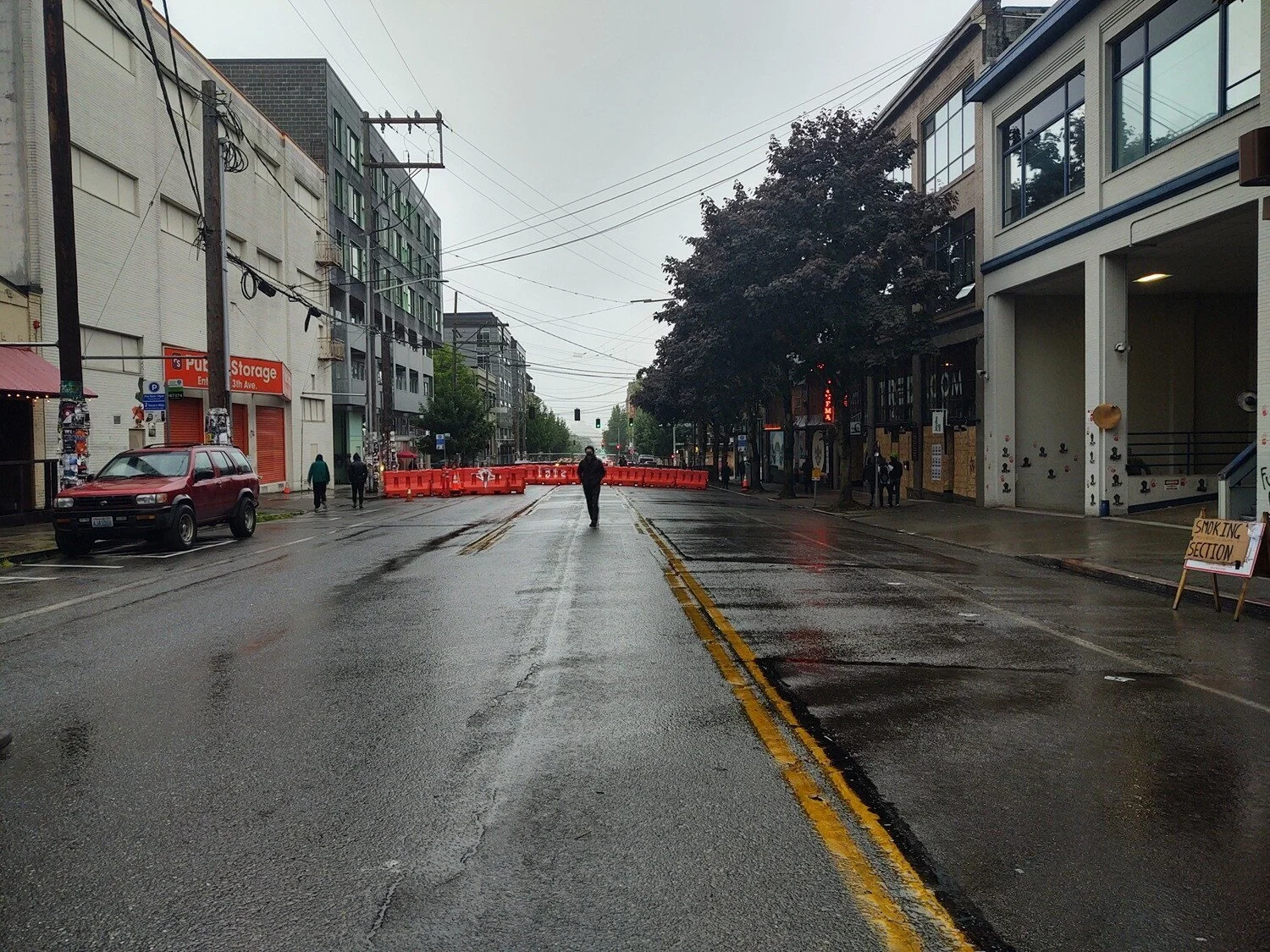 A person walks down the middle of a wet, empty street. A row of orange barricades are seen behind them.