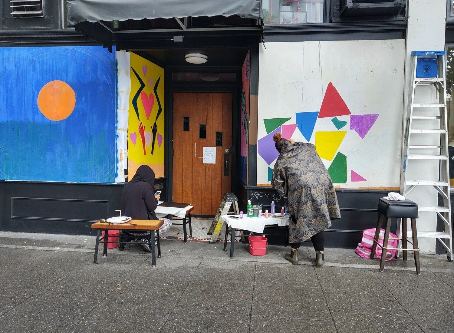 Art supplies sit on small tables as a hooded person, kneeling, looks at a phone and a person wearing a poncho paints shapes.