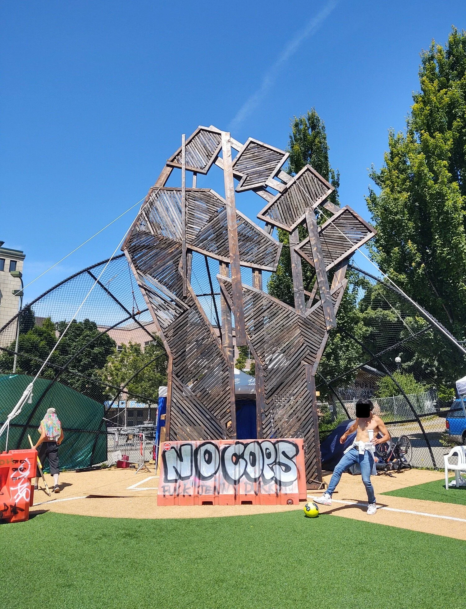 Two shirtless people. One kicks a yellow ball. One carries a stick. An orange barricade in front of statue reads “no cops”.