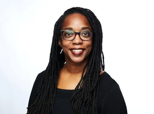 Tarika Powell poses for a professional photo wearing a black shirt, with long dreadlocks, in front of a white background.
