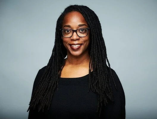 Tarika Powell poses for a professional photo wearing a black shirt, with long dreadlocks, in front of a blue background.